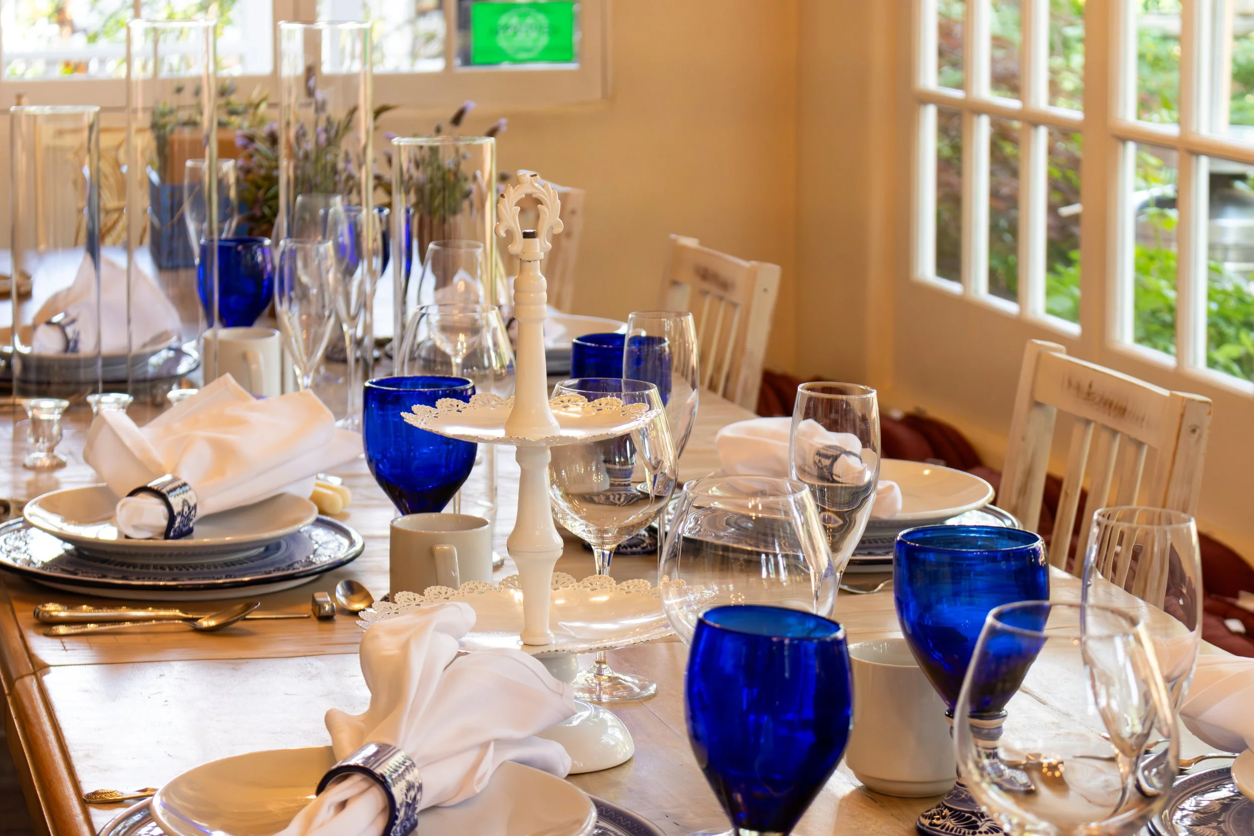 A dining table set for a formal meal with white napkins, blue glasses, and elegant tableware in a bright room with large windows and greenery outside.