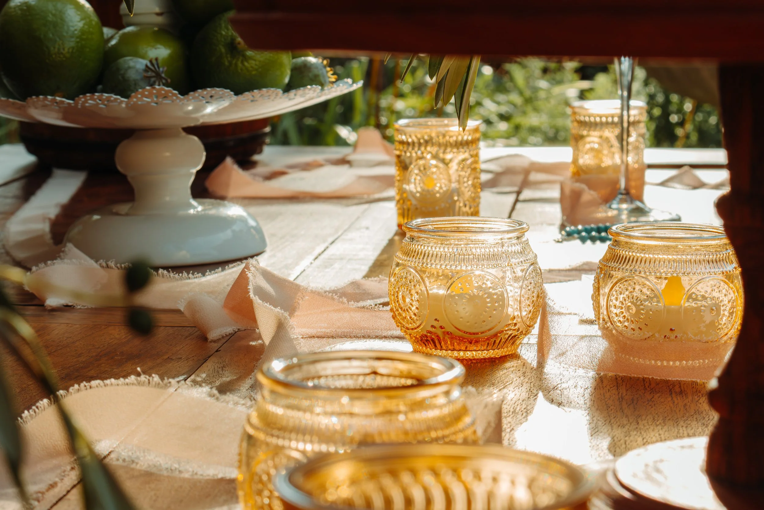 A rustic outdoor table decorated with amber glass candle holders, a porcelain cake stand with green limes, and a white lace-like table runner, illuminated by natural sunlight.