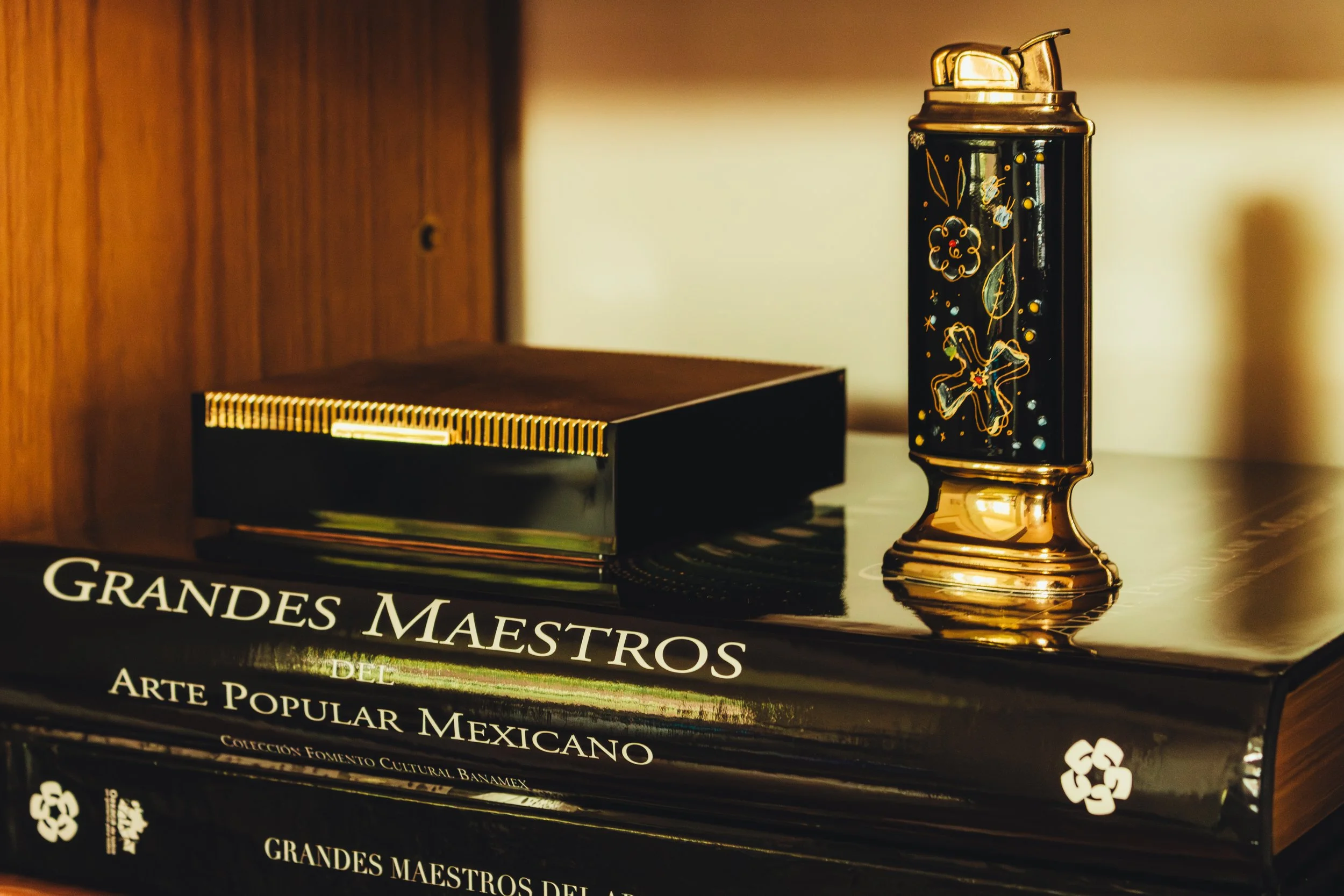 A book titled 'Grandes Maestros del Arte Popular Mexicano' with a black cover and gold accents, a lacquered box, and a decorative black and gold lighter or container with floral and fairy motifs on a wooden surface.