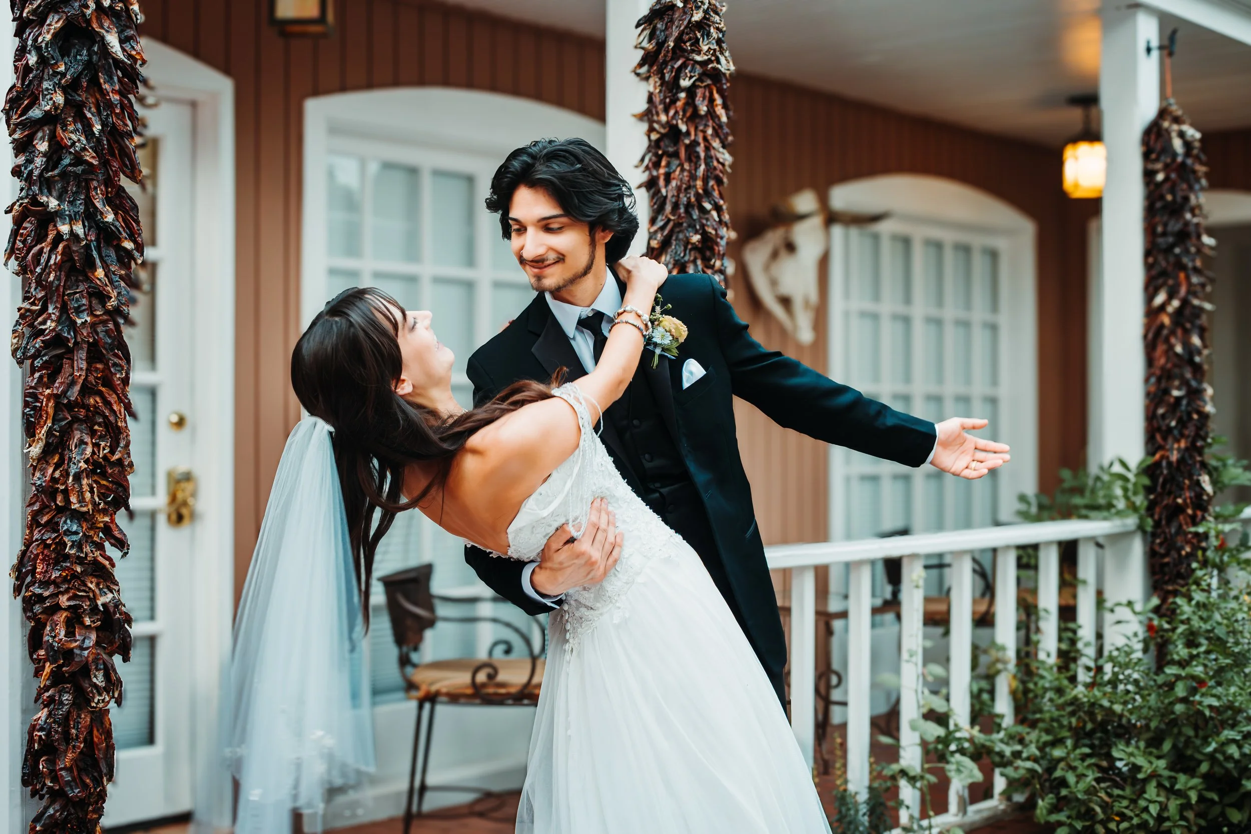 A groom in a black suit dancing with a bride in a white wedding dress on a porch with brown walls and white trim, decorated with dried chili ristras.