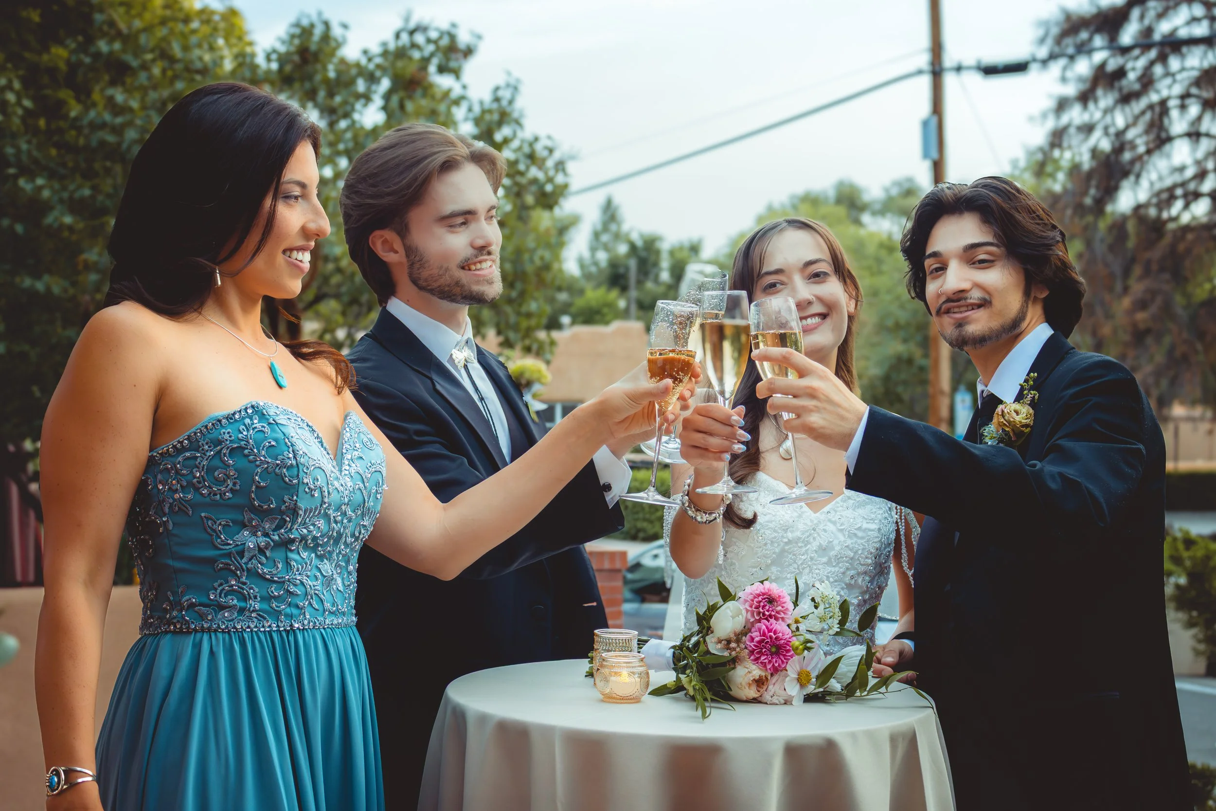 A group of five people celebrating at a wedding reception, raising glasses of champagne for a toast around a table with a bouquet and candles, outdoors with trees and power lines in the background.