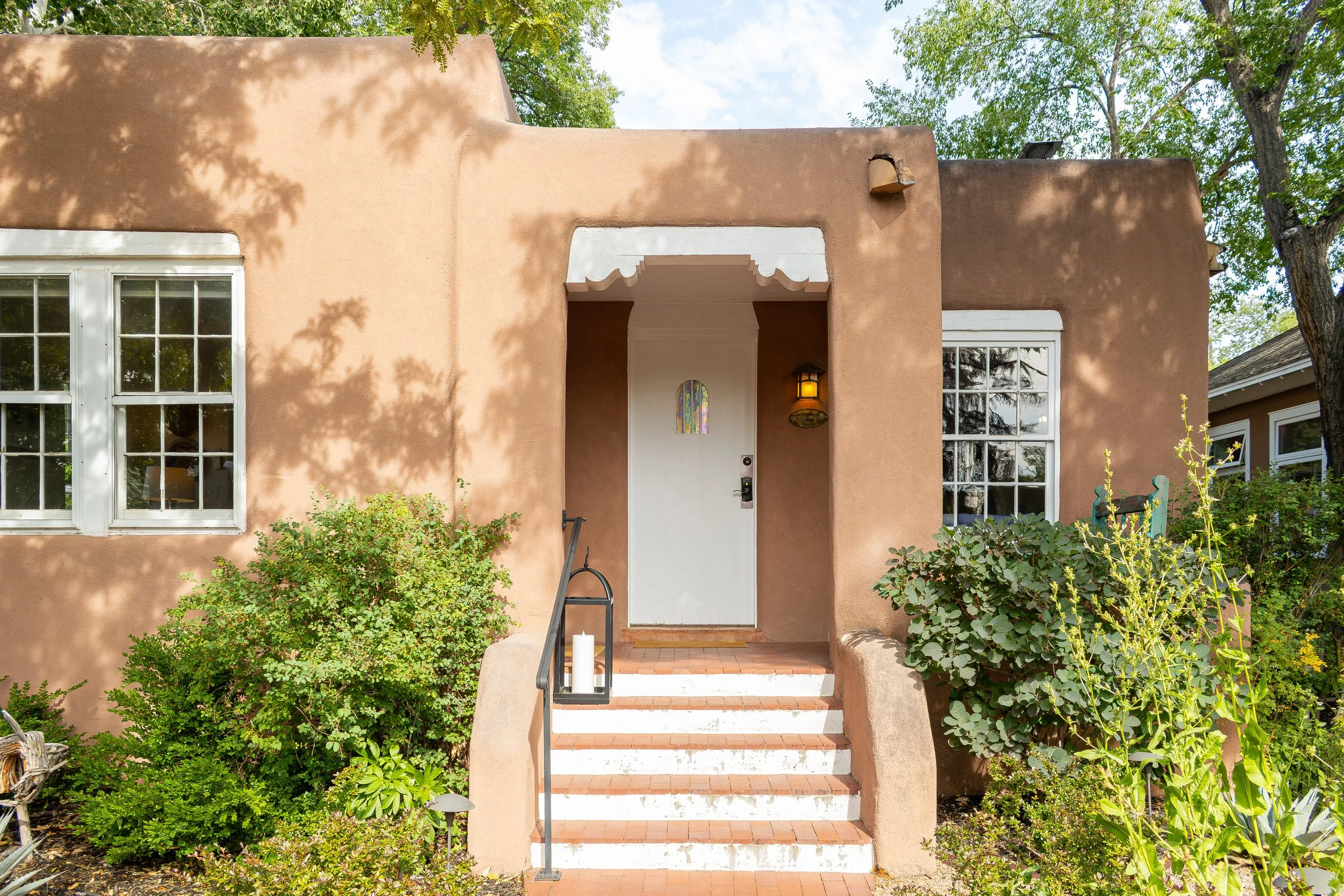 Adobe-style beige stucco house with white door, colorful stained glass window, and white-framed windows, surrounded by greenery and trees.