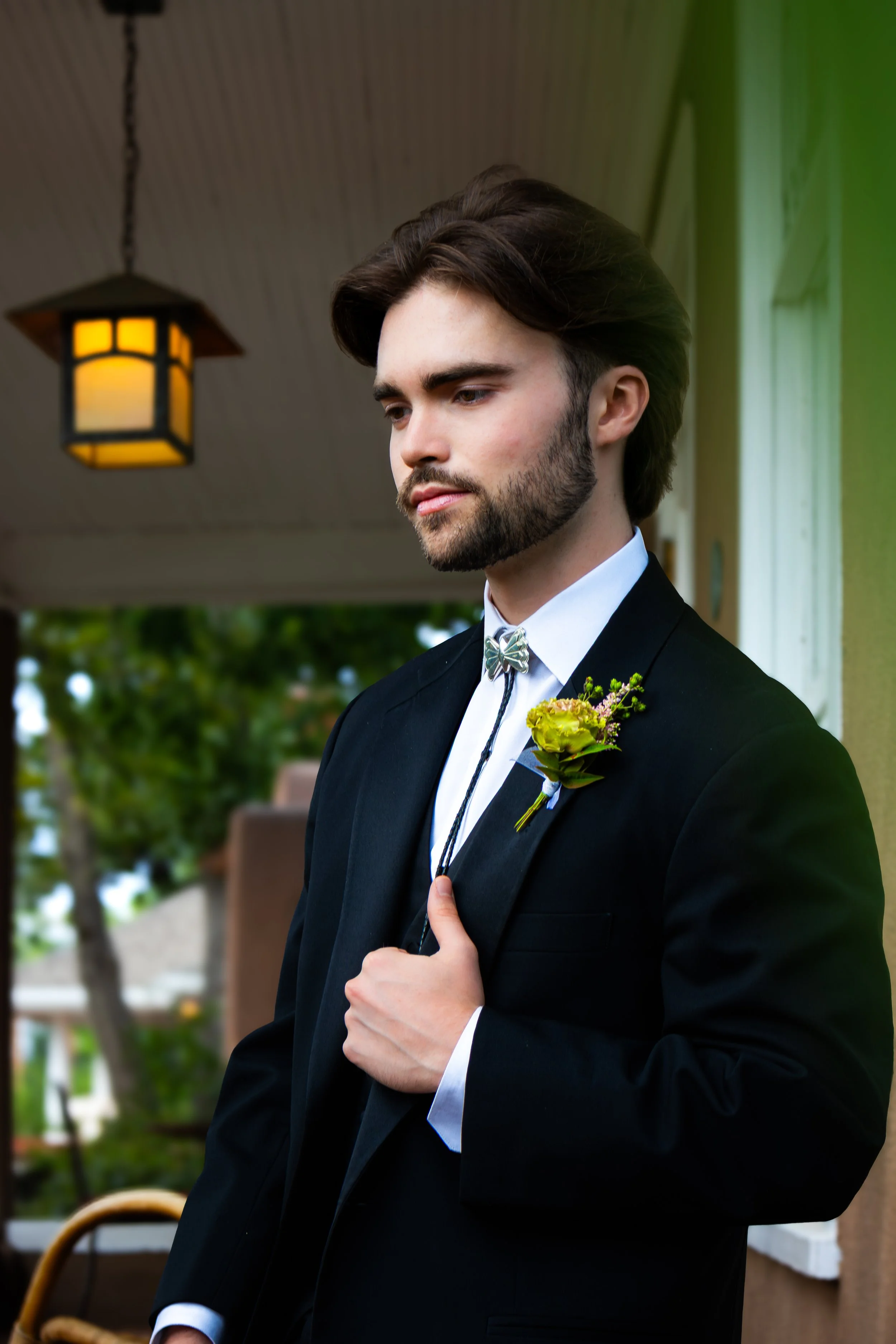 A young man with a beard in a black suit and white shirt, holding his suit lapel with his right hand, standing outdoors during daytime with trees in the background and a hanging light fixture above him.