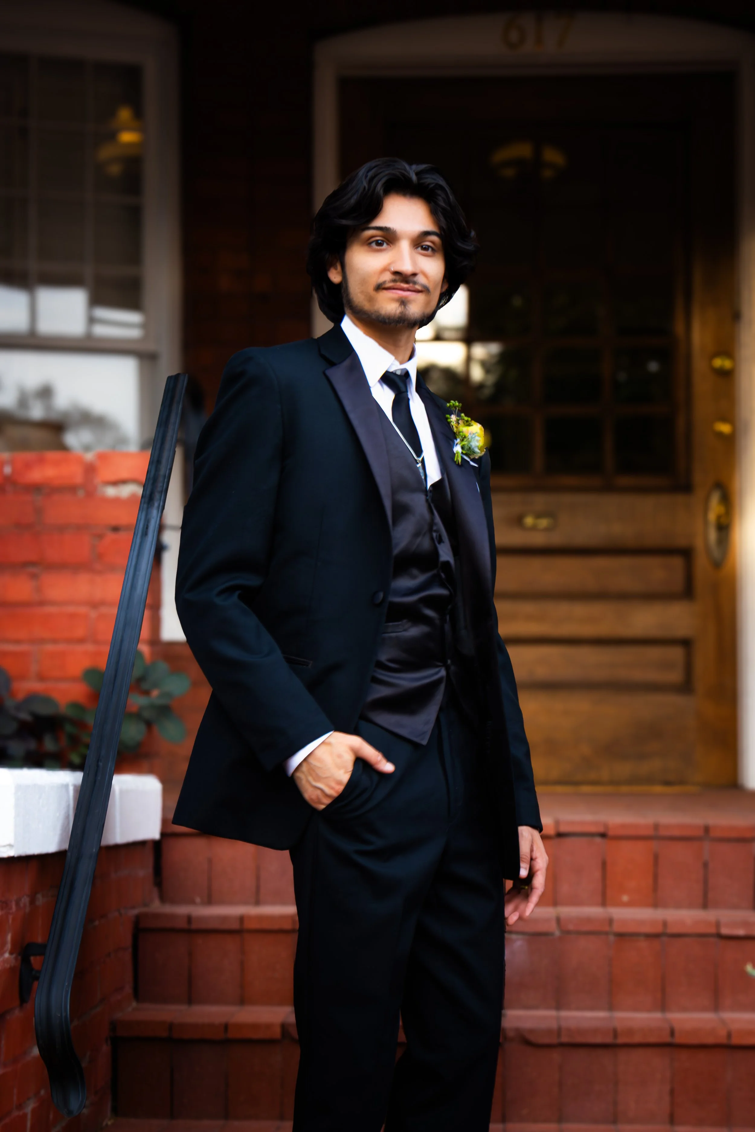 Young man in a black suit and tie standing on front steps of a house, with a boutonniere on his lapel, outside.