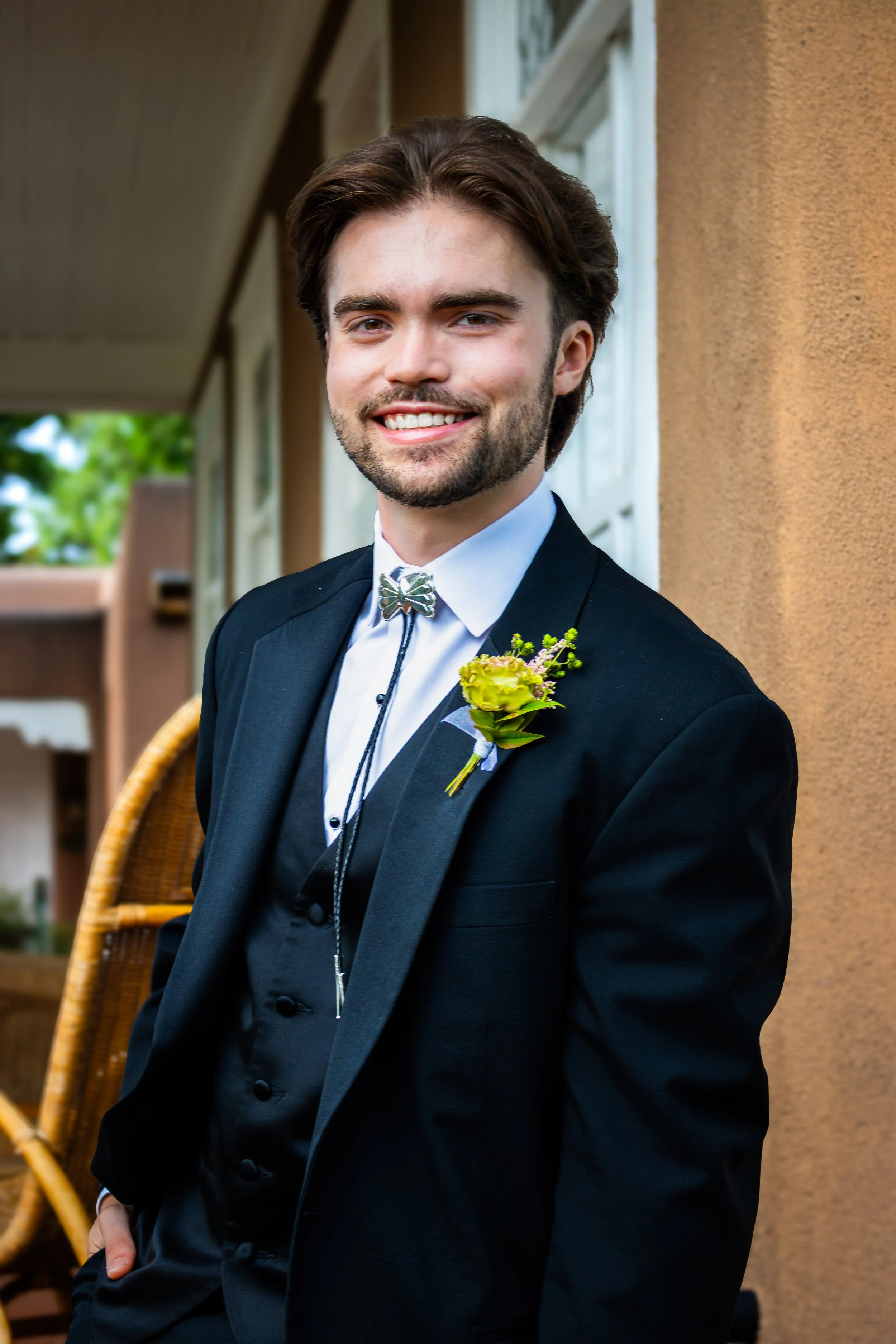 A young man in a suit and boutonnière smiling and posing outdoors.