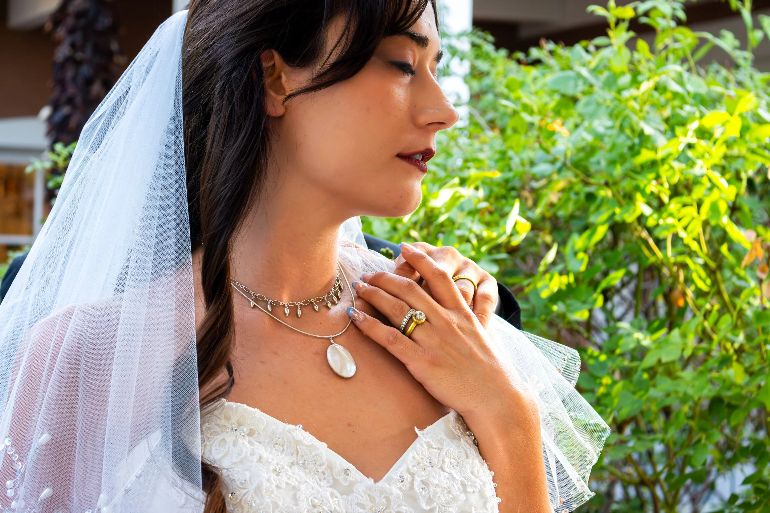 Close-up of a woman in a wedding dress with her eyes closed, holding her hand to her chest, wearing a veil and jewelry, outdoors with green foliage in the background.