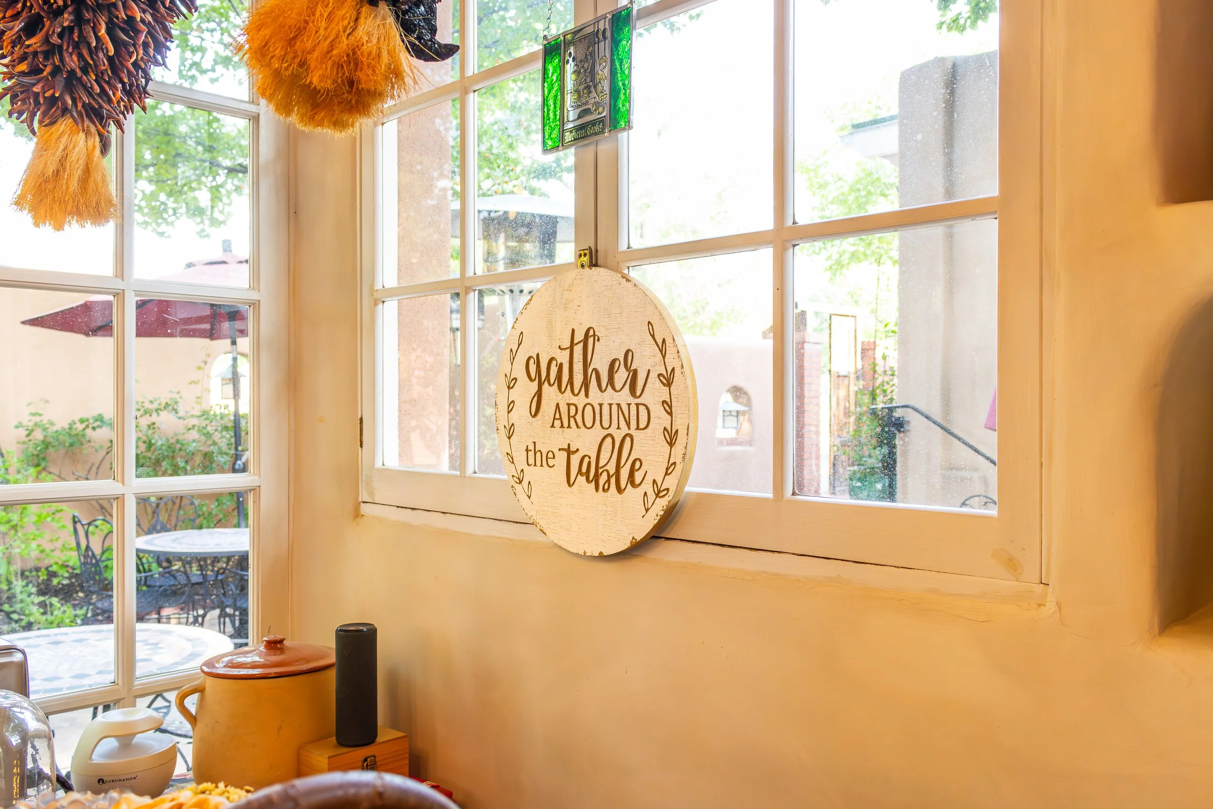 Interior of a cozy room with a large window showing an outdoor patio with tables and umbrellas. A decorative hanging sign reads 'Gather around the table.' Various hanging decorations and a small brown pot are visible on a surface below the window.