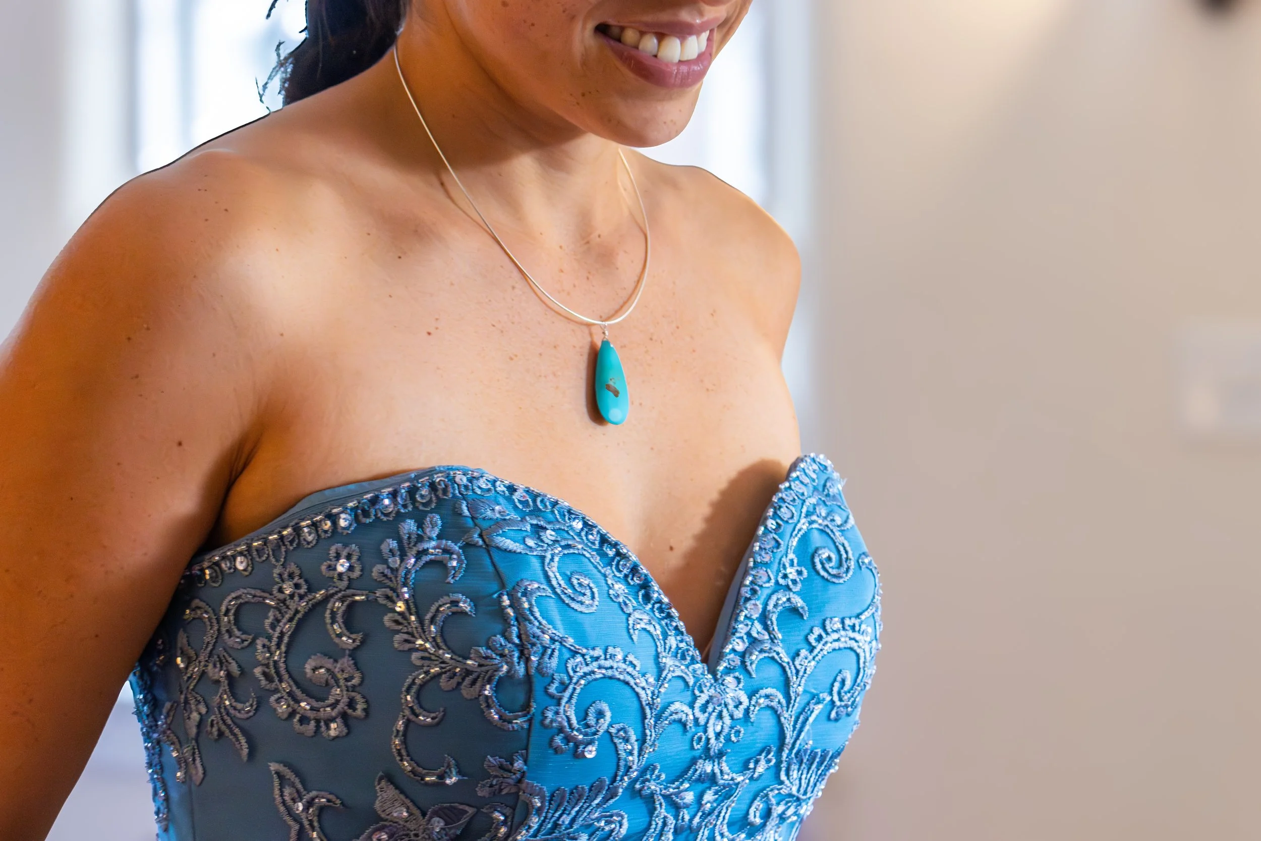 A woman wearing a strapless, ornate blue dress with detailed embroidery and beading, smiling and wearing a turquoise pendant necklace.