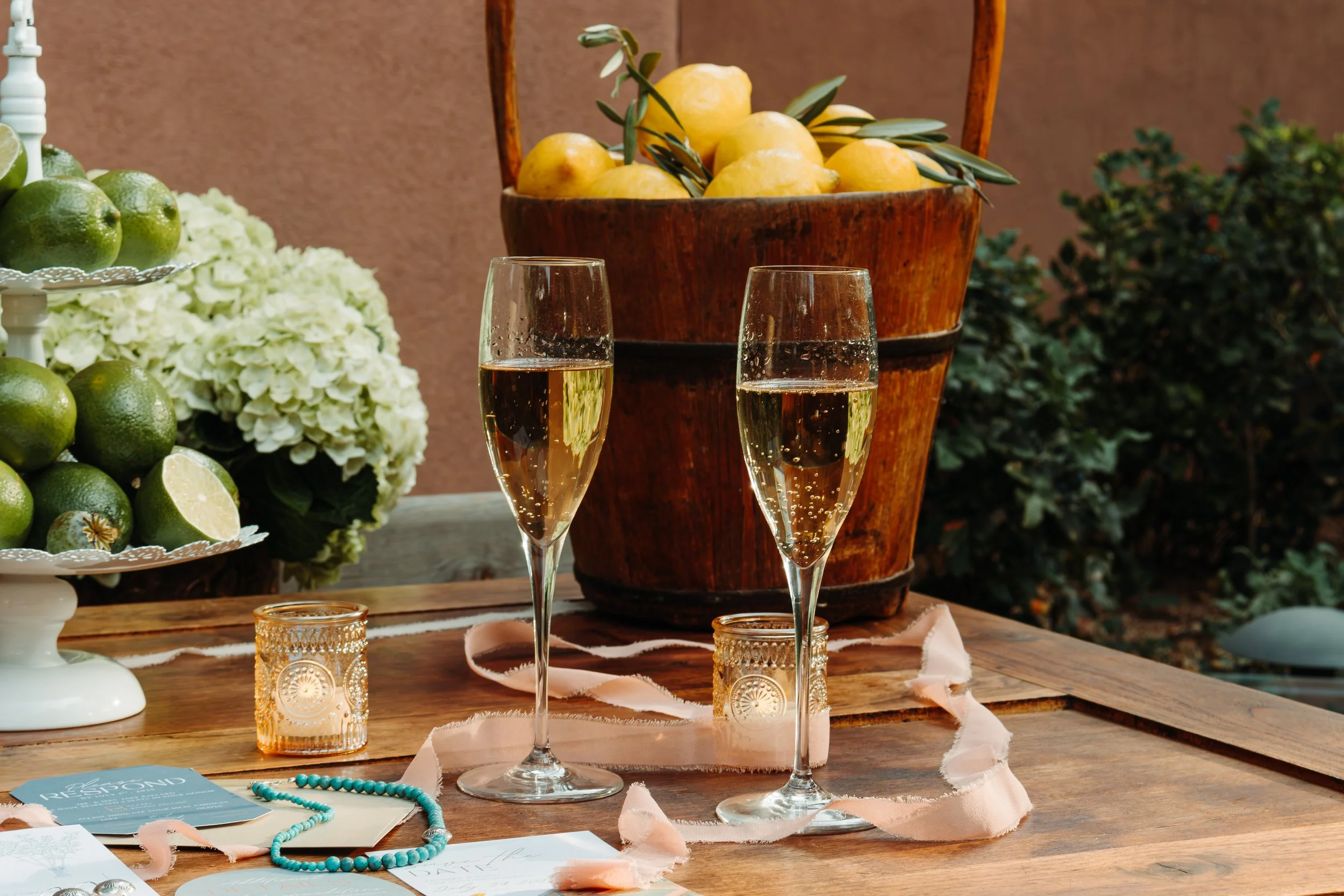 Two glasses of champagne on a wooden table, surrounded by candles, a turquoise beaded necklace, and paper with text, with a basket of lemons and a plate of limes with a lemon half in the background.