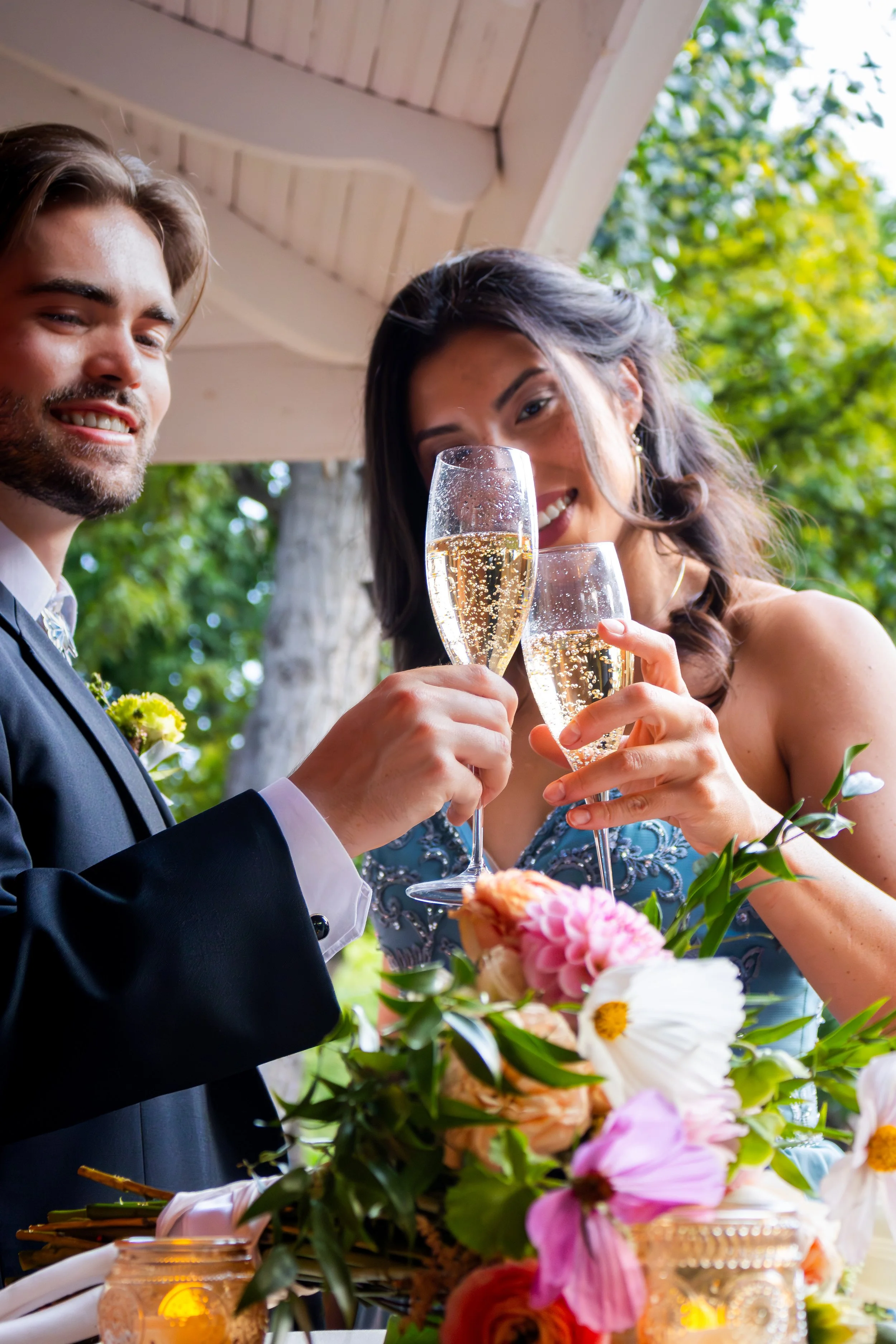 A man and woman dressed in formal attire clink champagne glasses during a wedding celebration outdoors, with a floral arrangement and candles on the table.