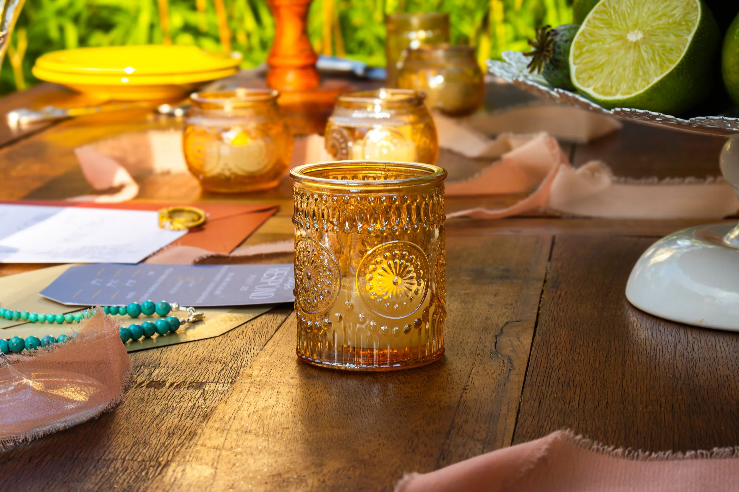 Decorative amber glass candle holder on a wooden table with other candle holders, lime halves, and jewelry around it, set outdoors with natural green background.