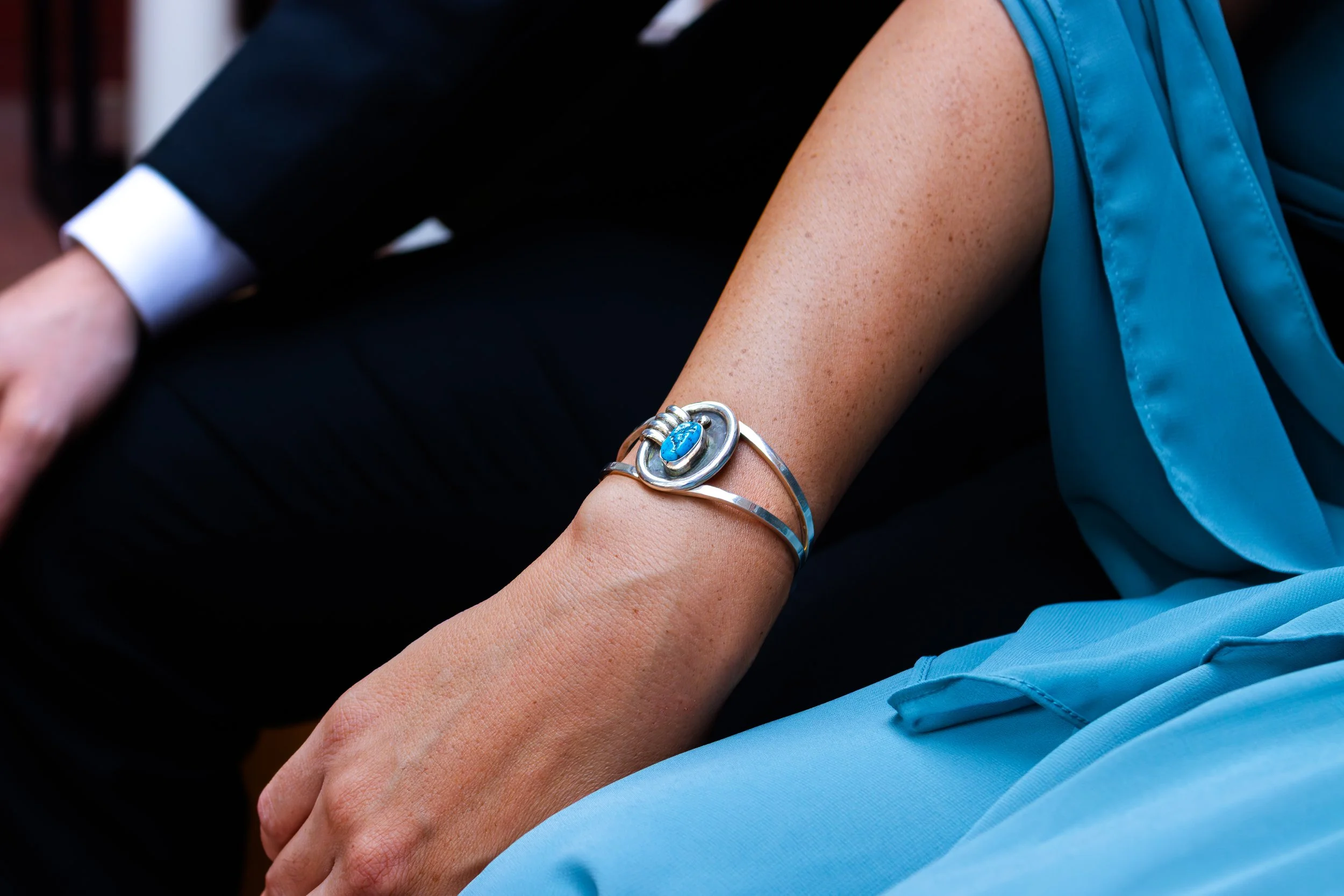 Close-up of a person's arm wearing a silver bracelet with turquoise and silver details, with a blue dress or blouse, and a person next to them in formal attire.