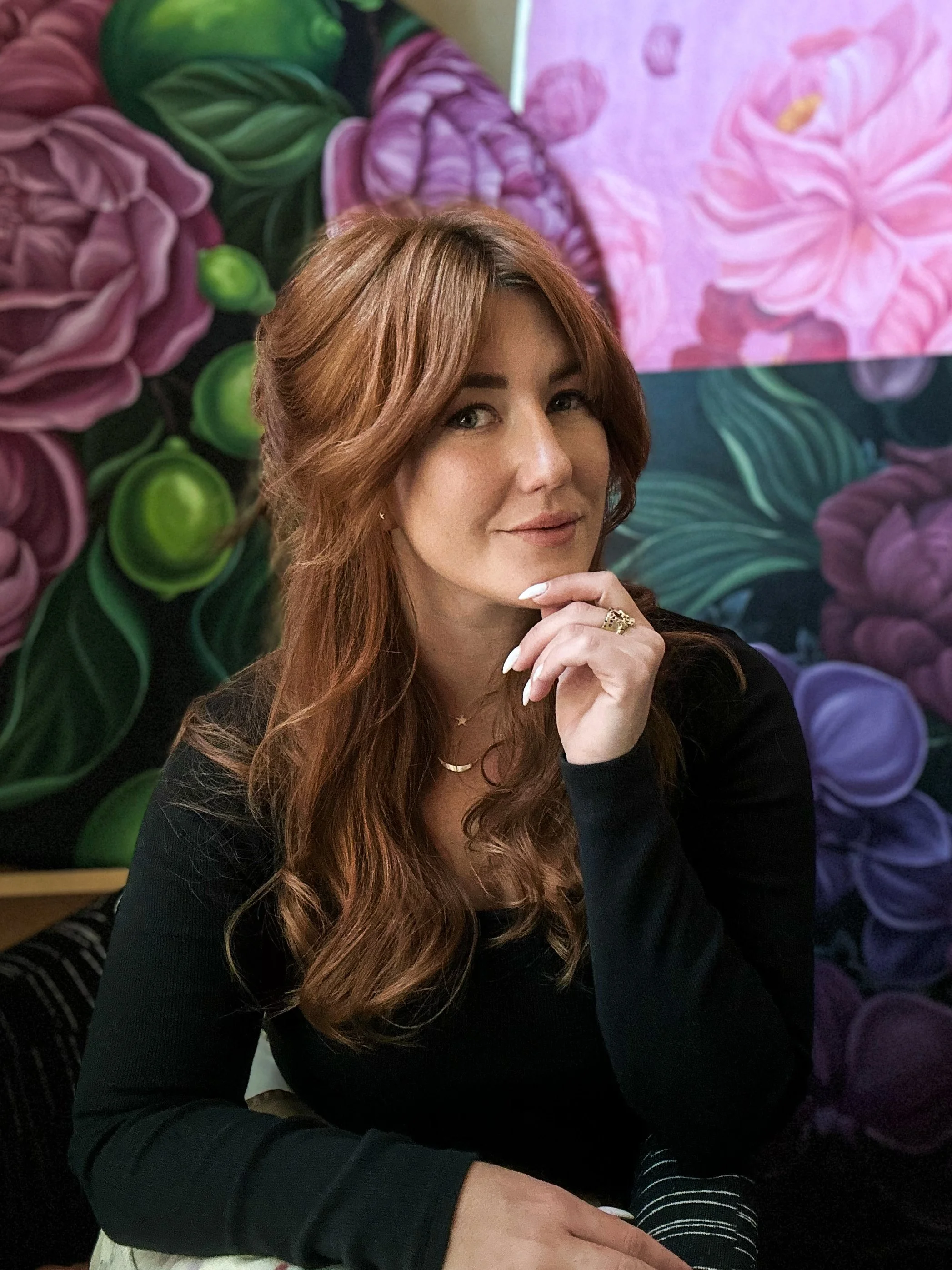 A woman with long wavy auburn hair in a black top, sitting with a hand near her chin and a faint smile, in front of a vibrant floral mural featuring pink, purple, and green flowers.