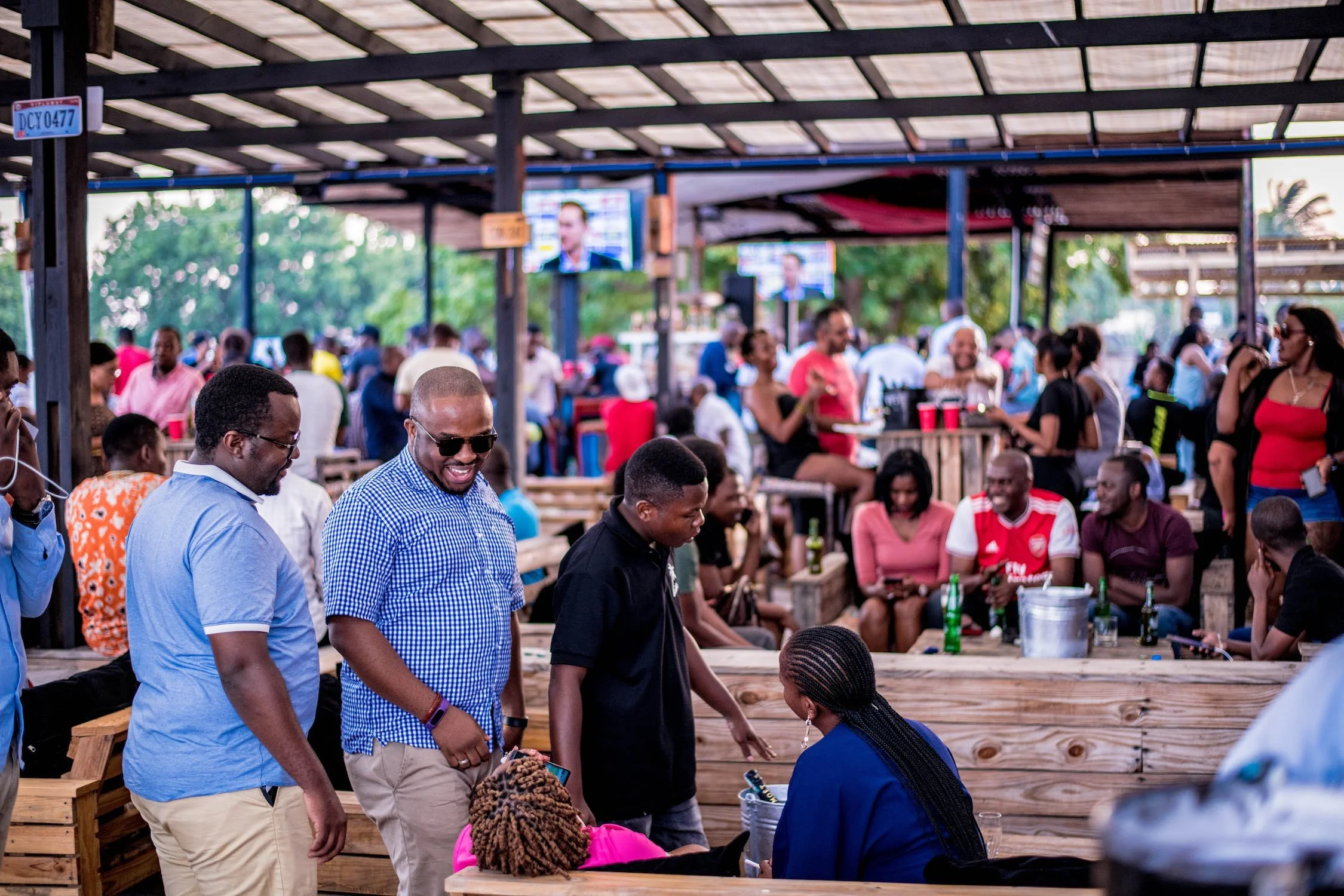 People enjoying a social gathering at an outdoor venue with wooden tables and a covered roof, some watching television screens, and others engaging in conversations, with a mix of men and women, drinking bottles of beer, and a relaxed atmosphere.