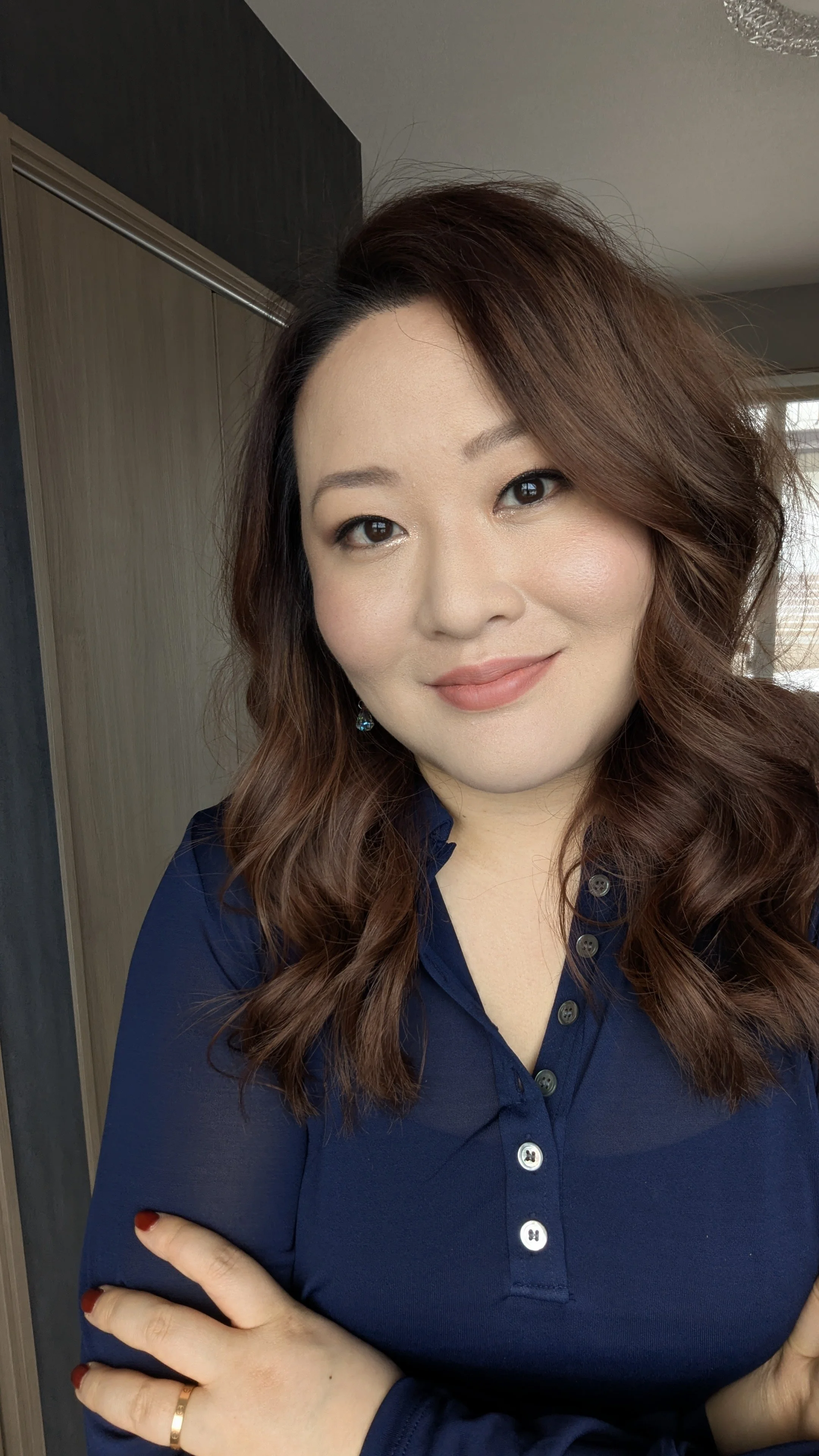 A woman with wavy auburn hair smiling, wearing a navy blue shirt with buttons, standing indoors near a closet.