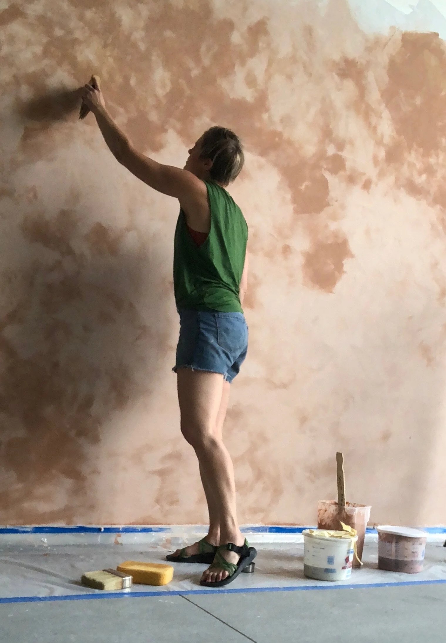 A woman painting a textured, peach-colored wall with a sponge brush, wearing a green tank top, denim shorts, and sandals, standing on a gray floor covered with painter's plastic and surrounded by buckets of paint and painting supplies.