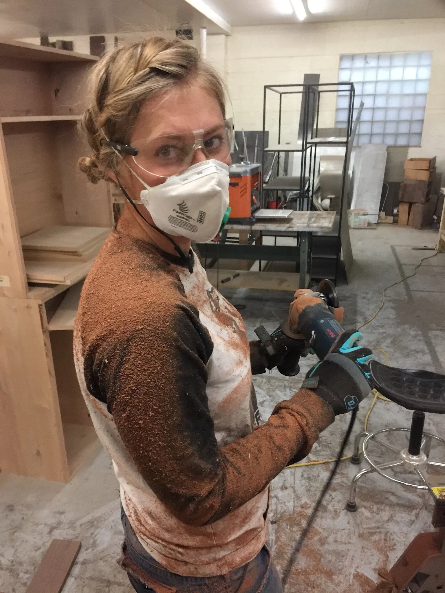 Woman working in a woodworking shop with safety glasses, a dust mask, and gloves, holding a handheld power sander.