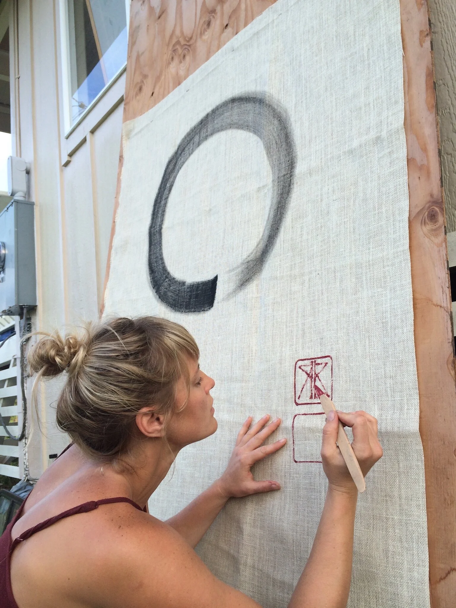 A woman with blonde hair tied up in a bun working on a large fabric canvas outdoors, drawing a black circular shape and red symbols with a red marker.