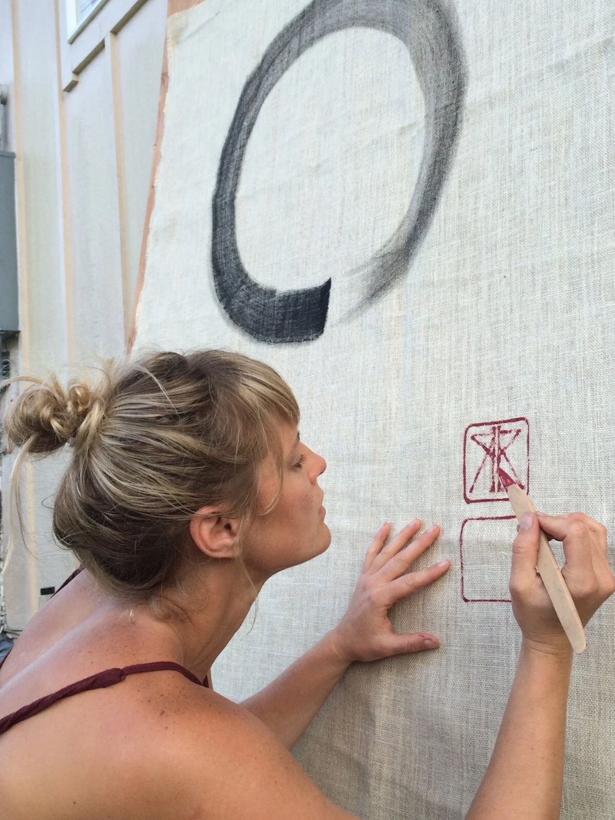 A woman with blonde hair tied up in a bun working on a large fabric canvas outdoors, drawing a black circular shape and red symbols with a red marker.