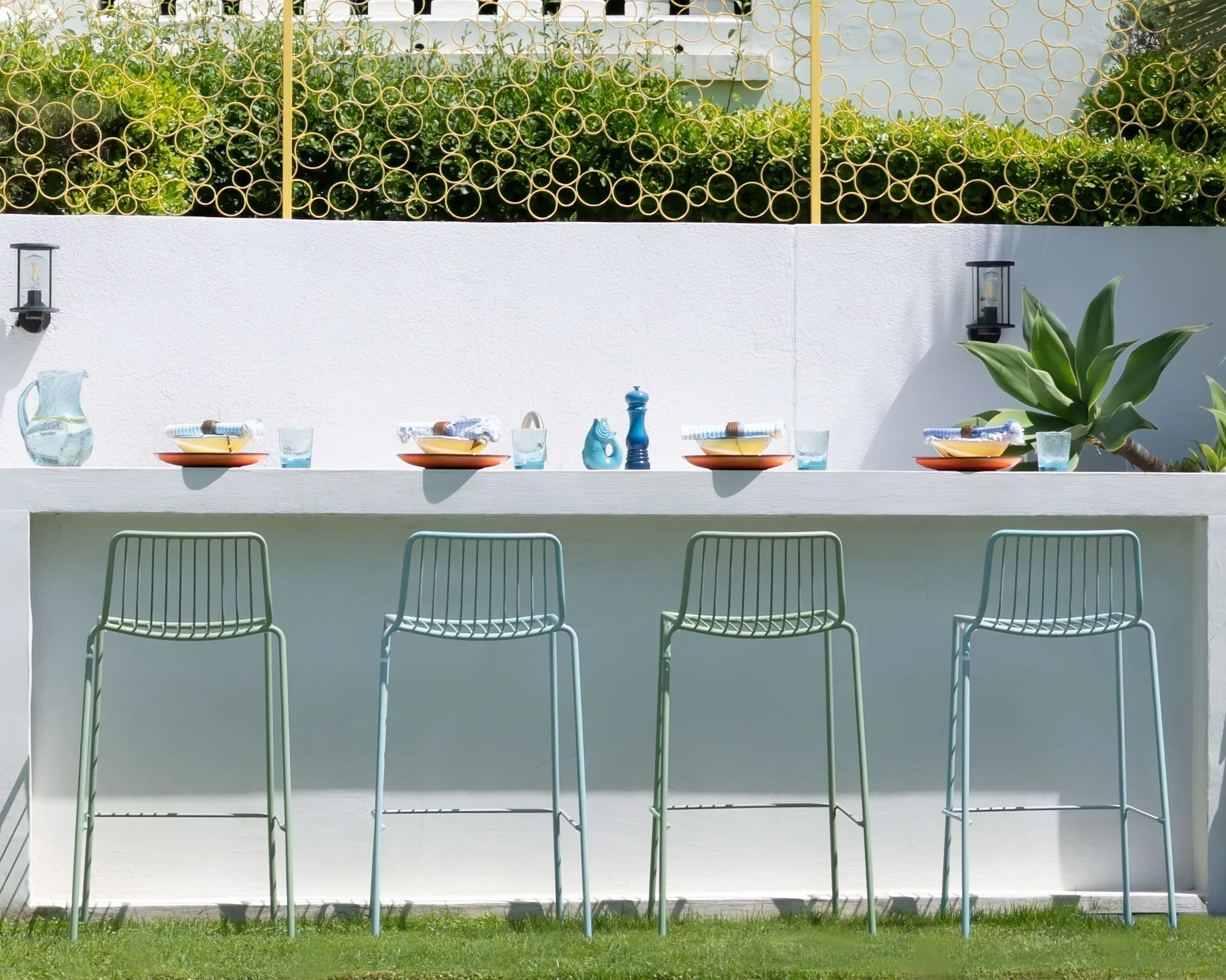 Outdoor dining setup with a white counter, four light green metal bar stools, colorful plates and glasses, a pitcher of water, napkins, and decorative items. A large green plant and a black wall-mounted lantern are in the background, with a decorative gold wire fence above.