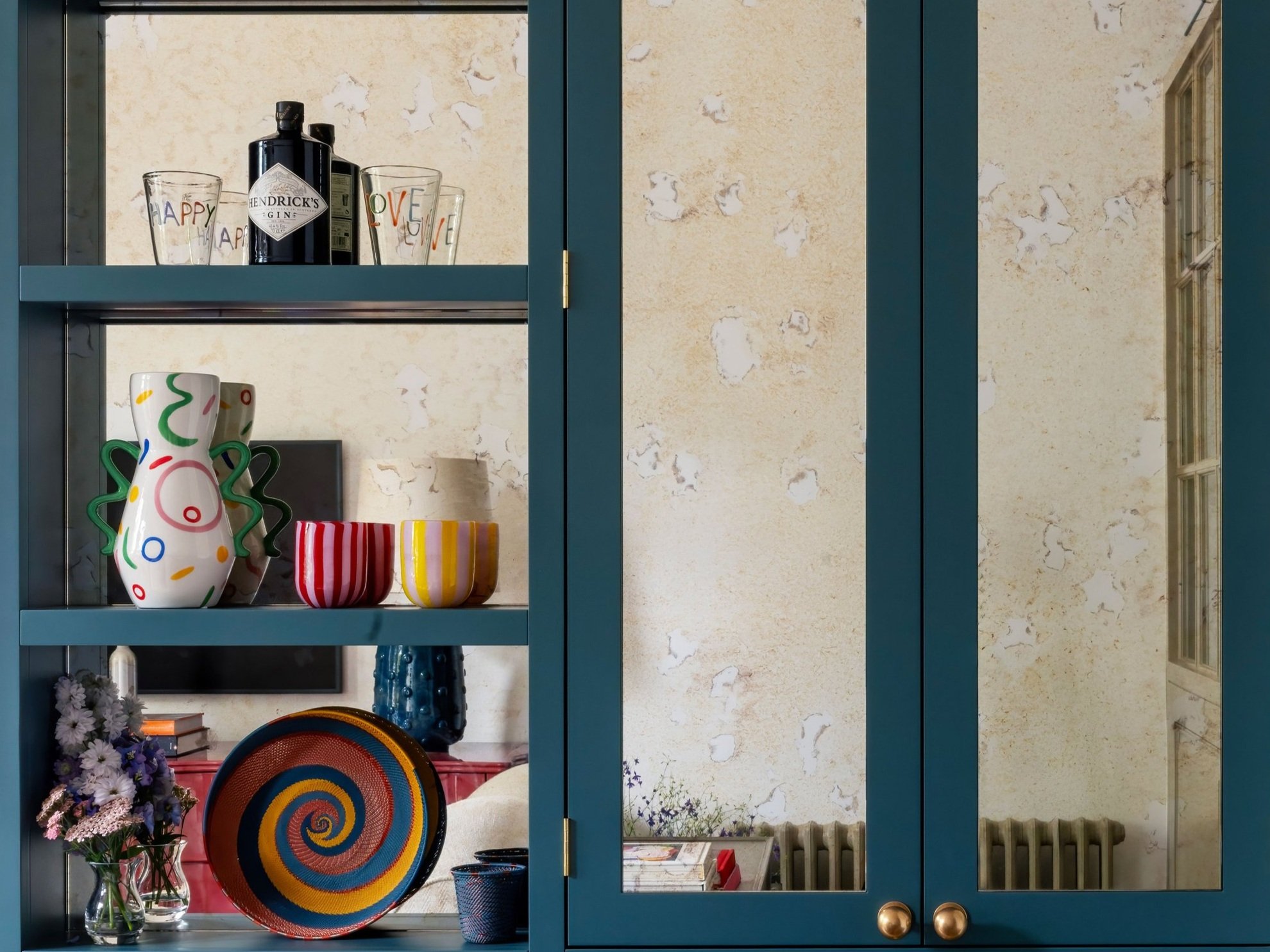 Decorative blue cabinet with colorful glassware, bottles, and ceramics, including striped cups, a multicolored swirl plate, and vases with flowers, against a textured beige wall.
