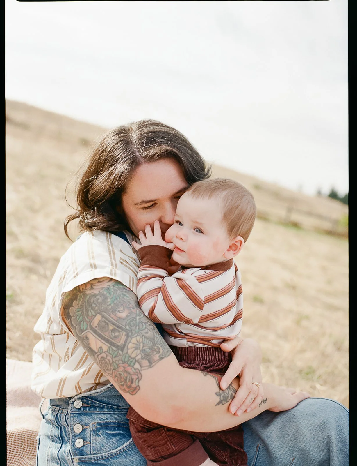 A woman with tattoos holding a young child outdoors on a grassy hill.