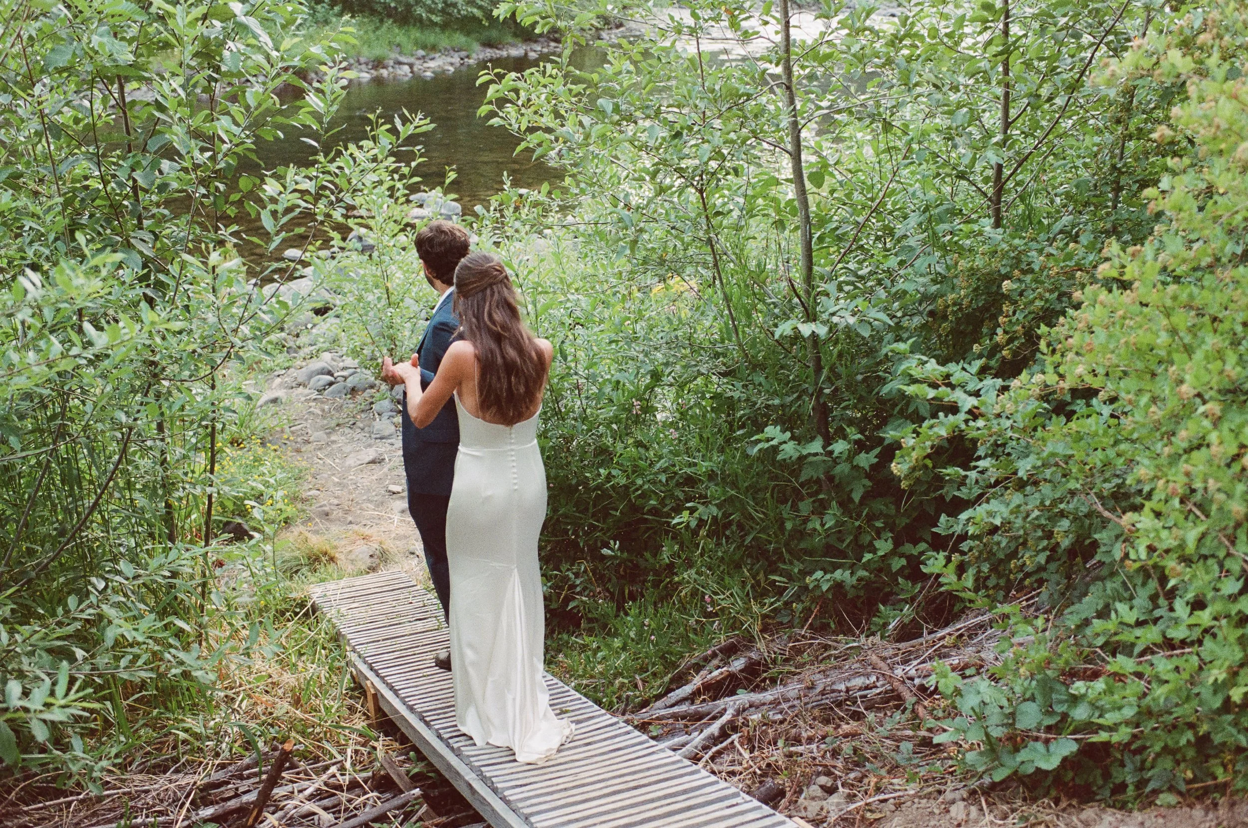 A newlywed couple stands on a small wooden bridge next to a stream, surrounded by lush green foliage.