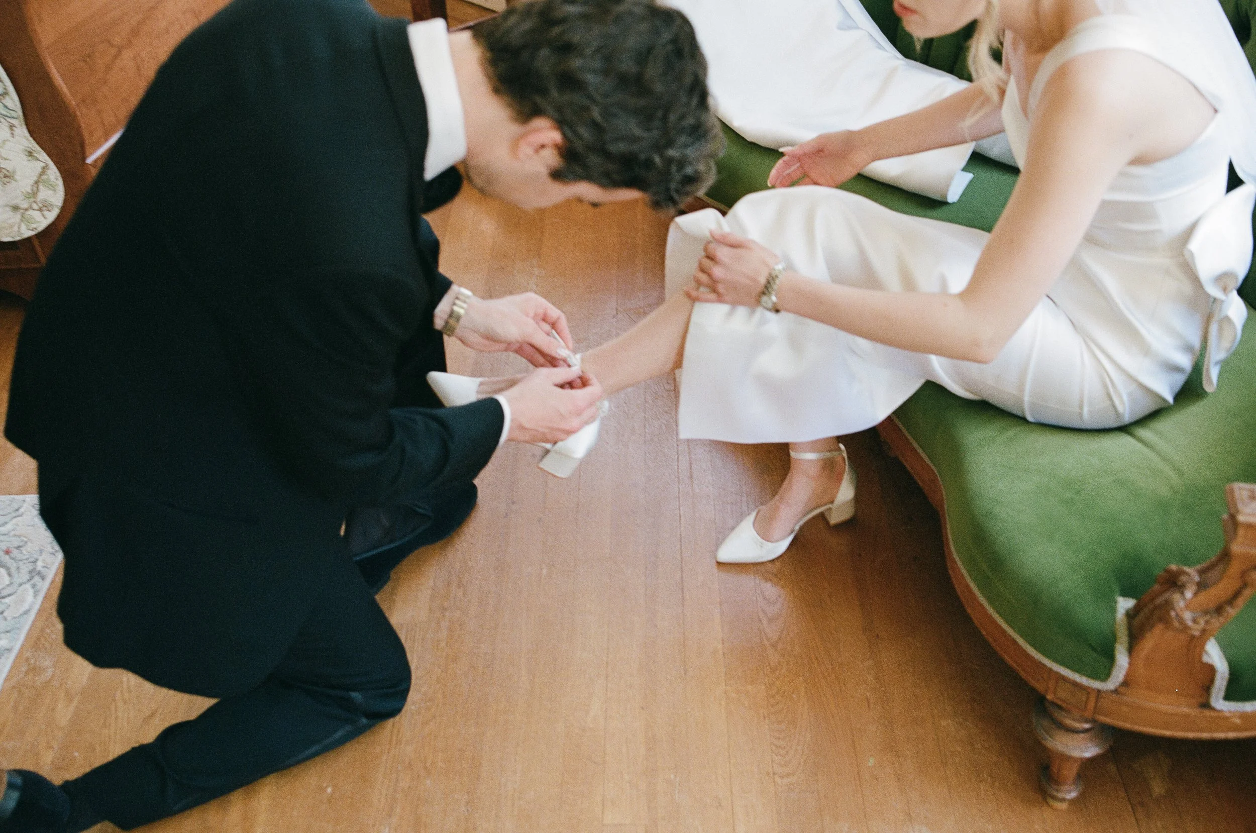 A man kneeling and fastening the shoes of a woman sitting on a green sofa, dressed in white, in a room with wooden flooring.