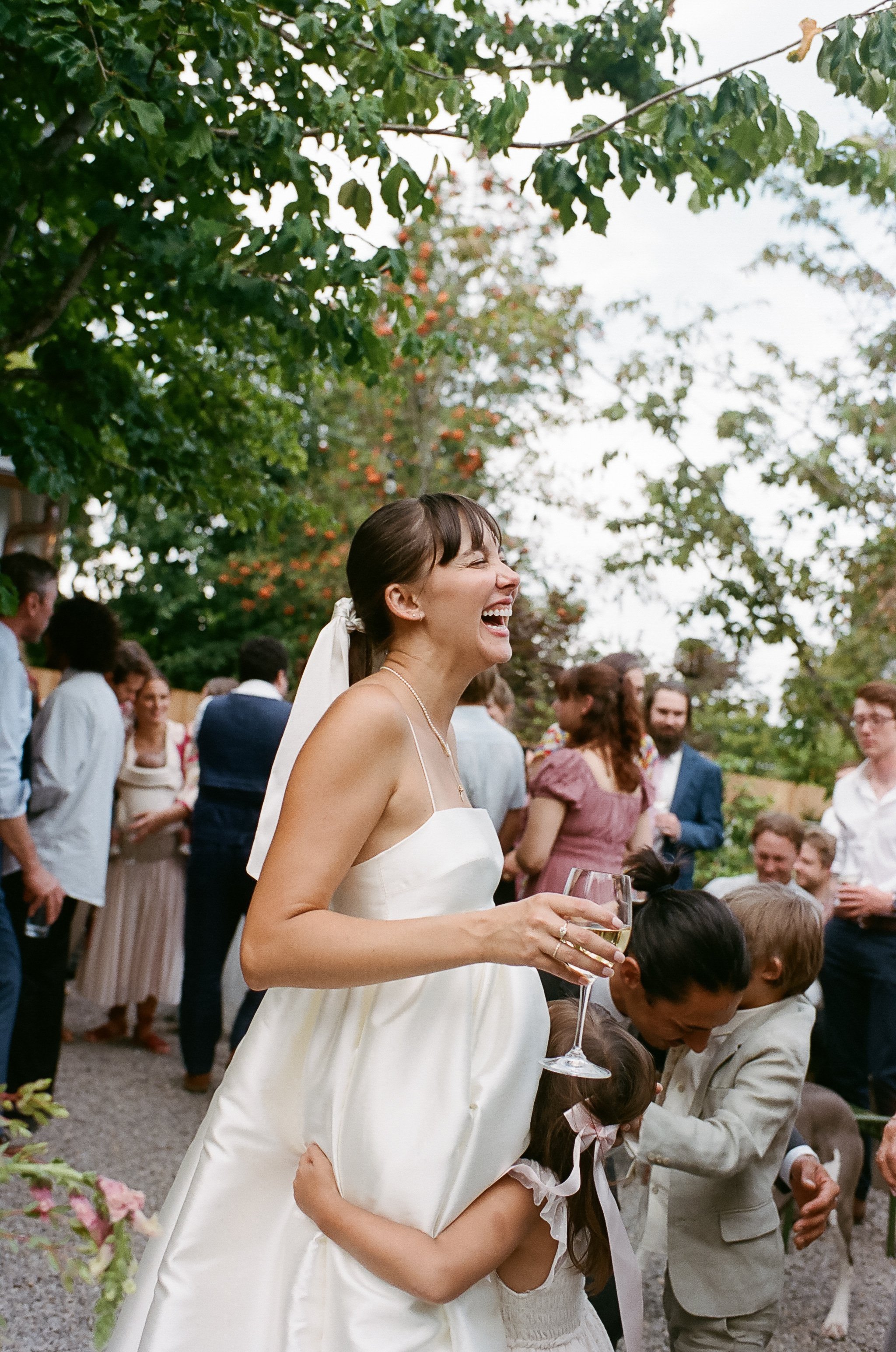 A woman in a white dress holding a glass of white wine, smiling joyfully at an outdoor celebration surrounded by children and adults under a leafy tree.
