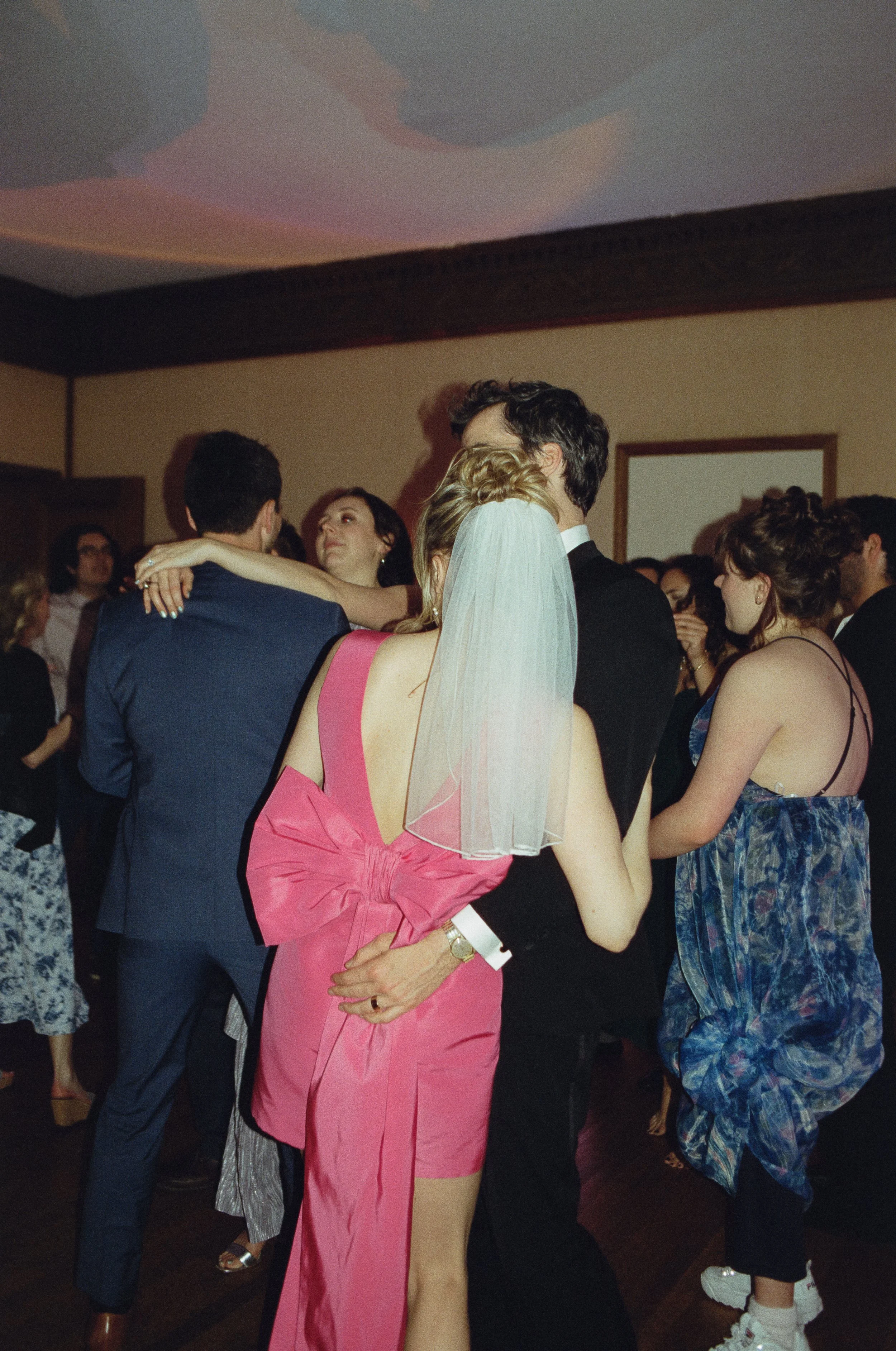 People dancing and socializing at an indoor celebration, with a woman in a pink dress and veil dancing with a man in a black suit, others in formal attire surrounding them.