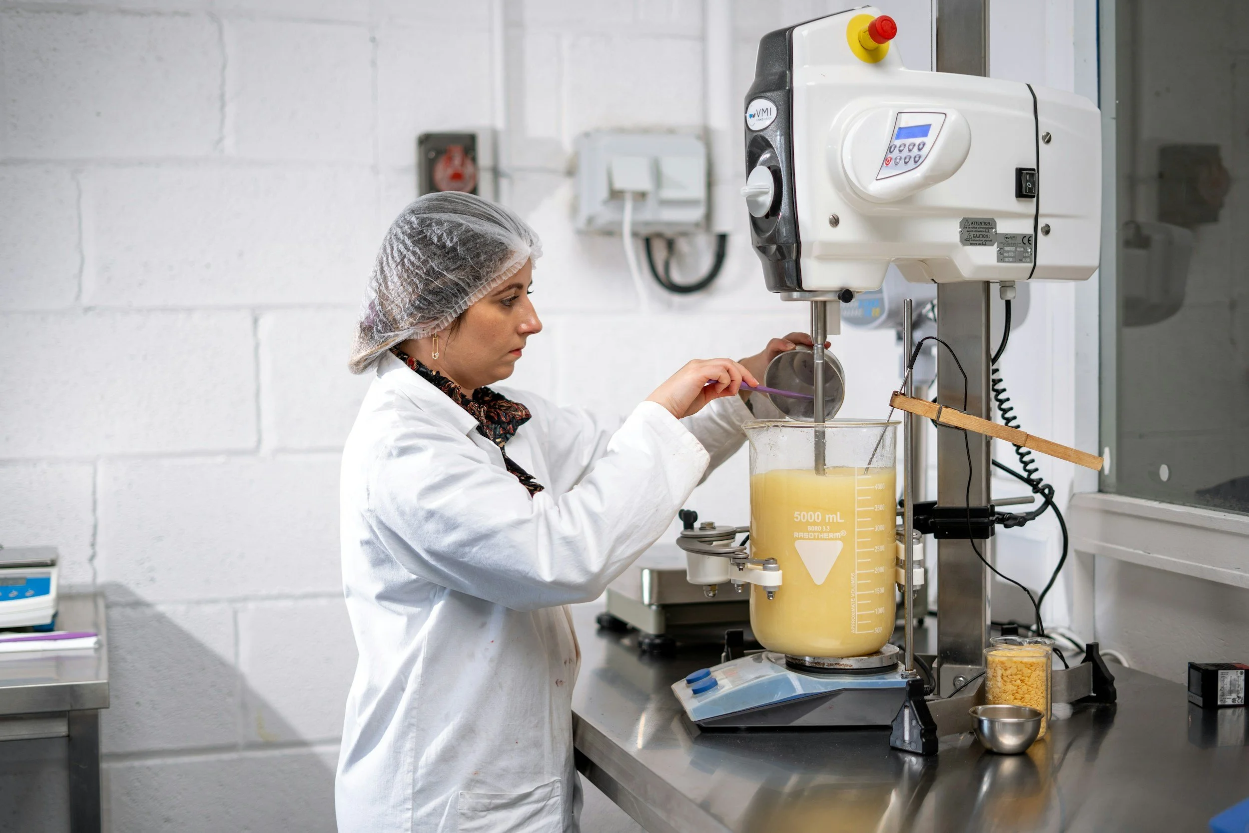 A woman wearing a hairnet and lab coat working with a large beaker containing a yellow liquid in a laboratory setting. She is operating a piece of scientific equipment with various controls.