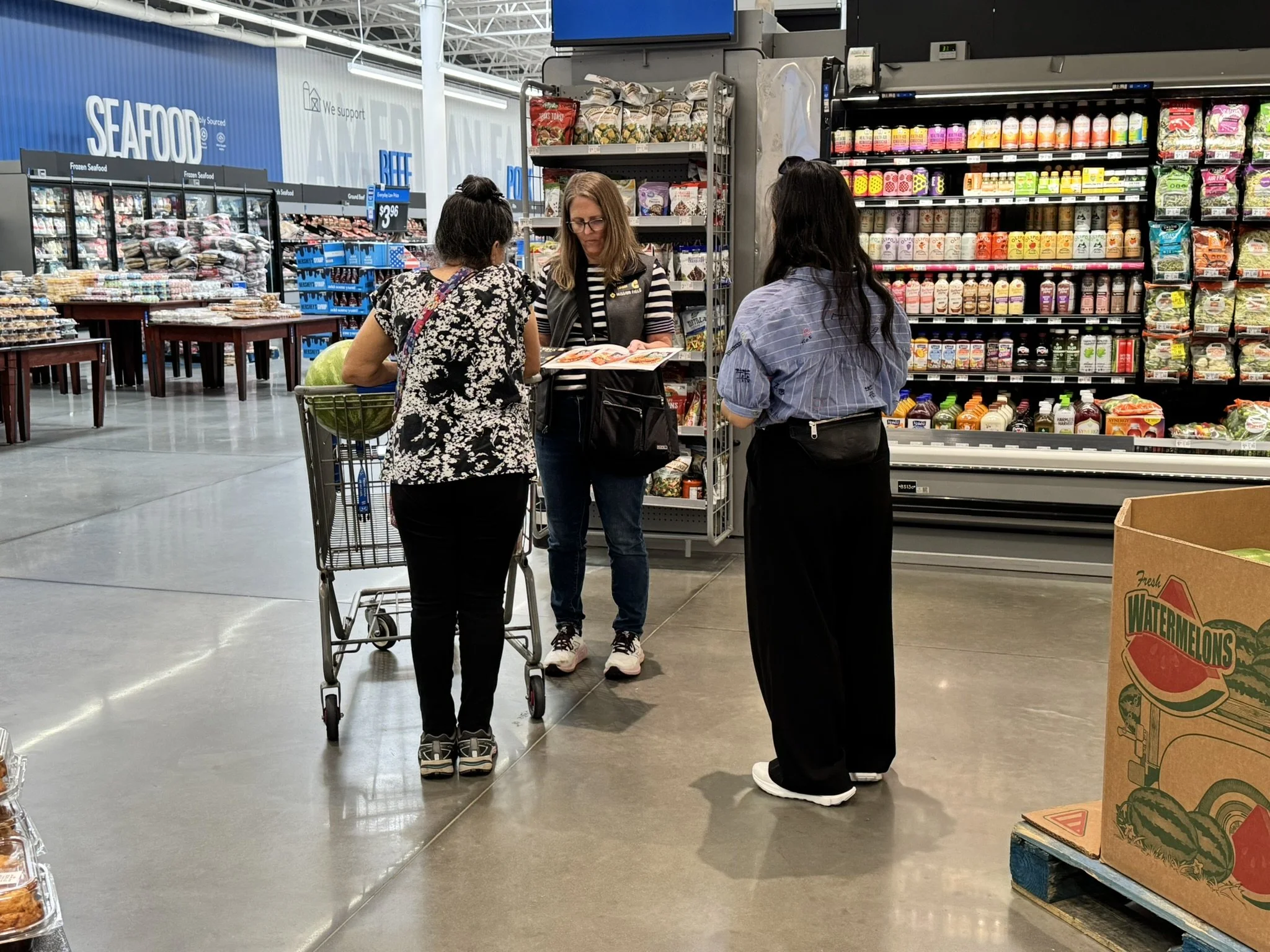 Three women are in a grocery store aisle. Two are standing behind a shopping cart, and one is interviewing them.