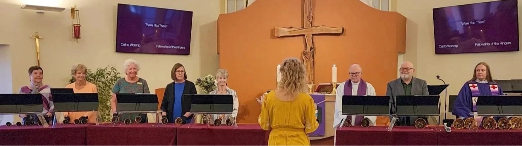 People standing behind a table inside a church, with a woman in a yellow dress facing them. The church has a cross on the wall, candles, and two TV screens displaying the words 'Were You There'.