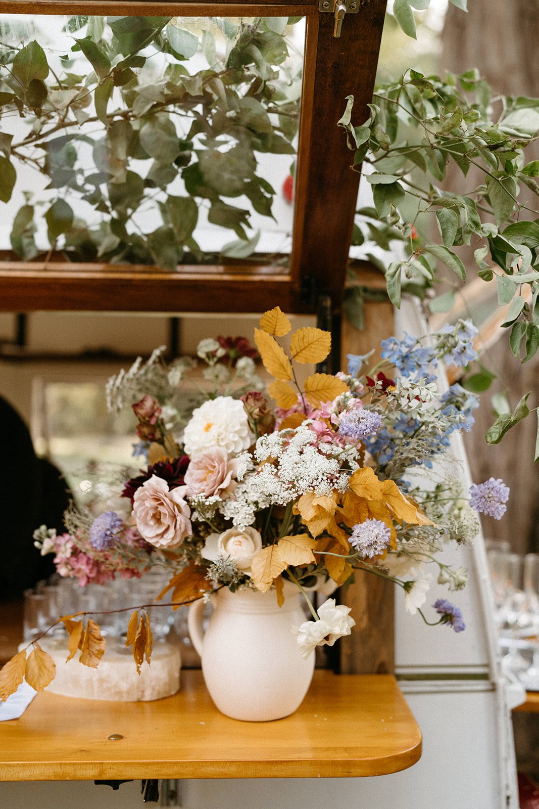 Colorful flower arrangement in white pitcher on a wooden surface, surrounded by green leaves and glassware.