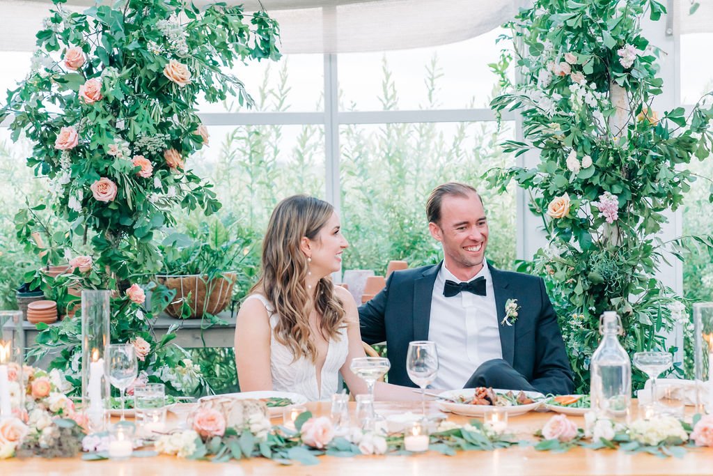 Bride and groom sitting at a decorated wedding table with floral arrangements.