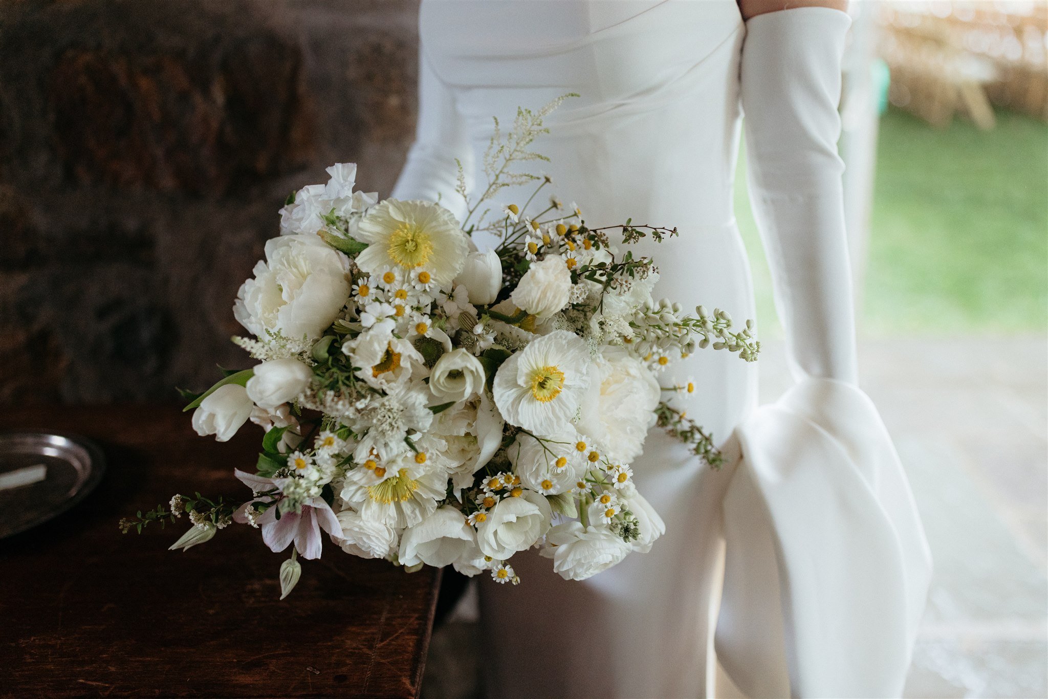 Bride holding white floral bouquet with daisies and greenery, wearing a white wedding dress and gloves.
