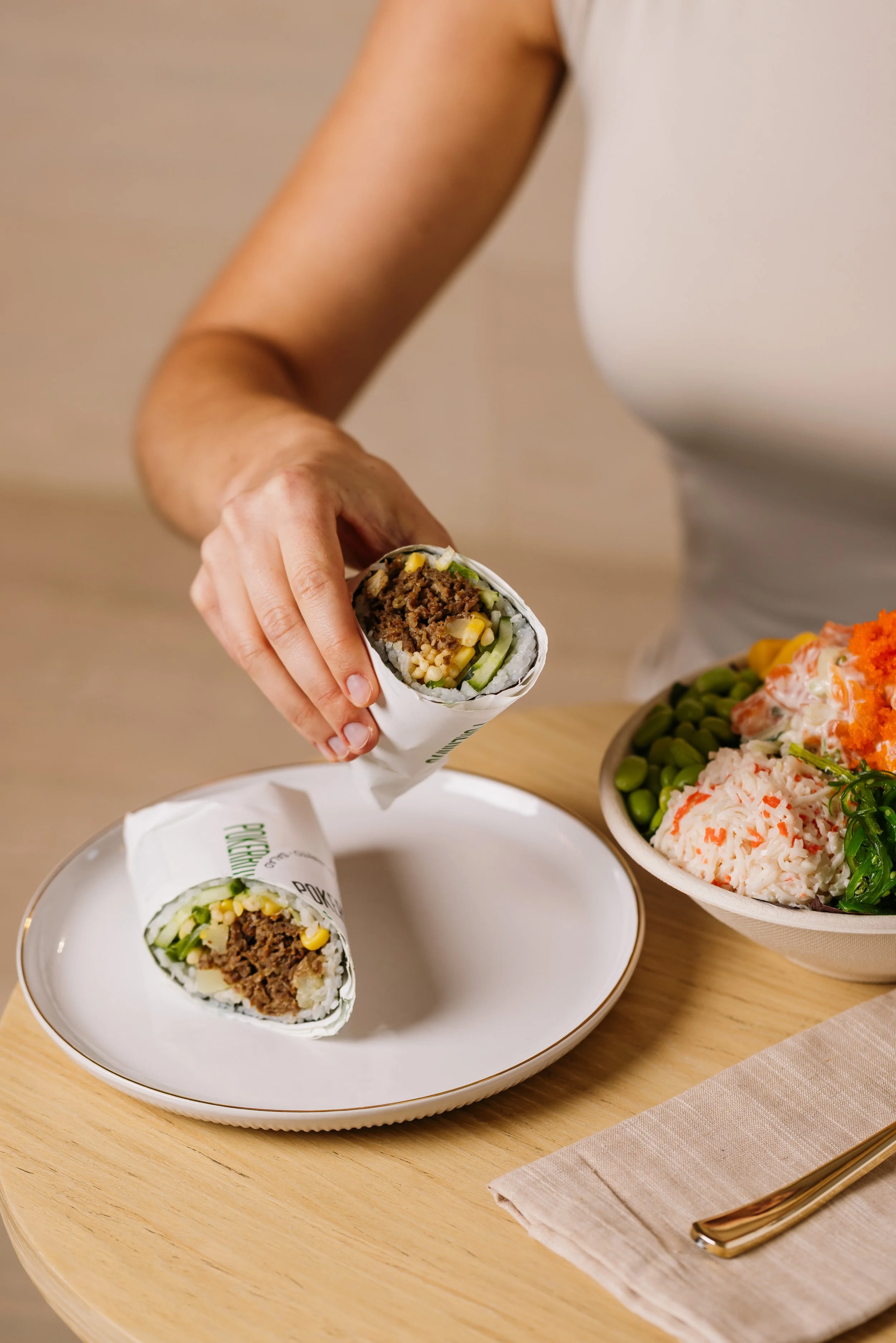 Close-up of a hand holding a sushi burrito filled with ground beef, corn, cucumbers, and rice, on a white plate with a second sushi burrito on a table nearby. A bowl of rice with imitation crab, green peas, and diced vegetables is beside the plate. A beige napkin and gold-colored fork are on the table.