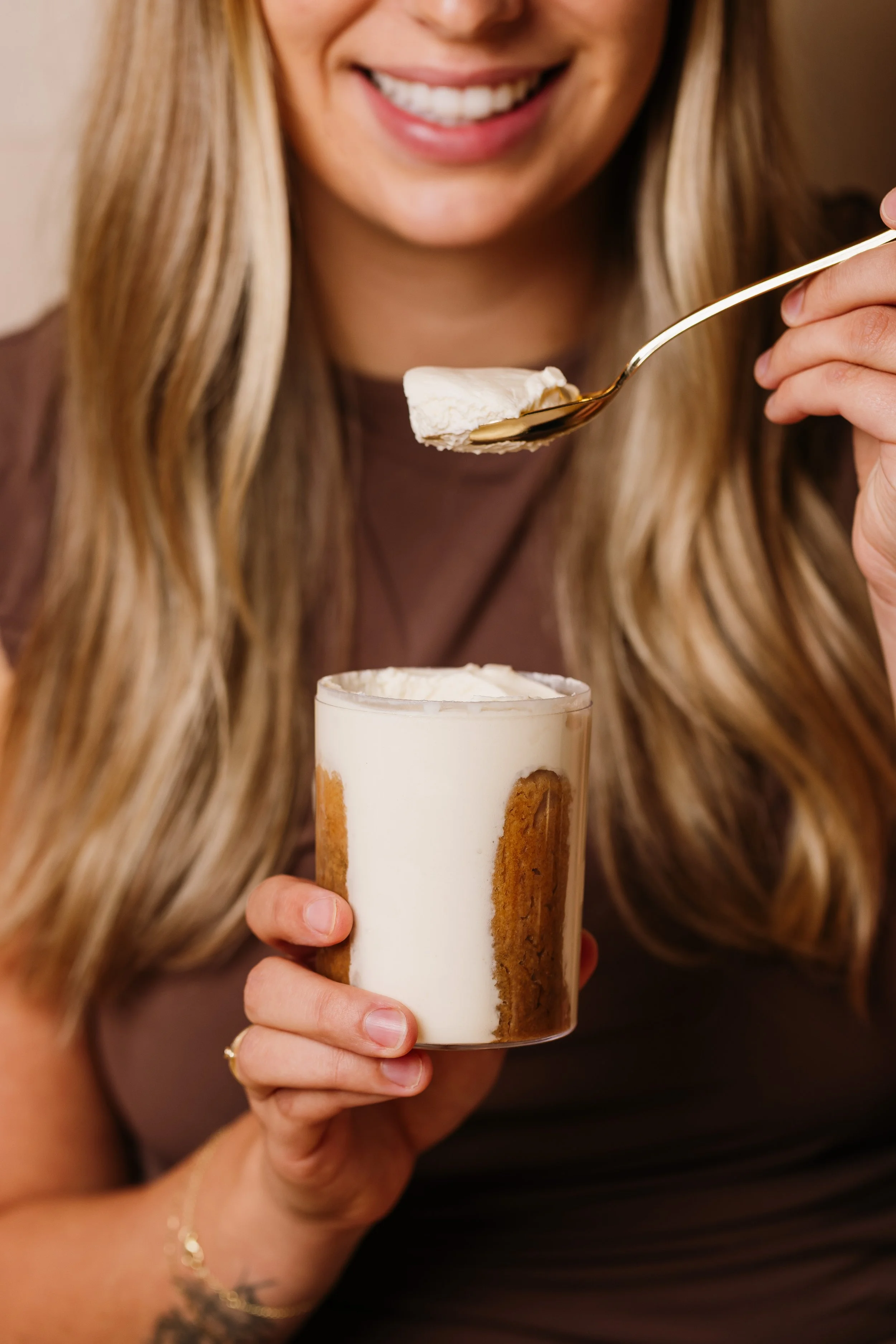 Close-up of a woman smiling, holding a glass of cookies and cream milkshake topped with whipped cream with a gold spoon.