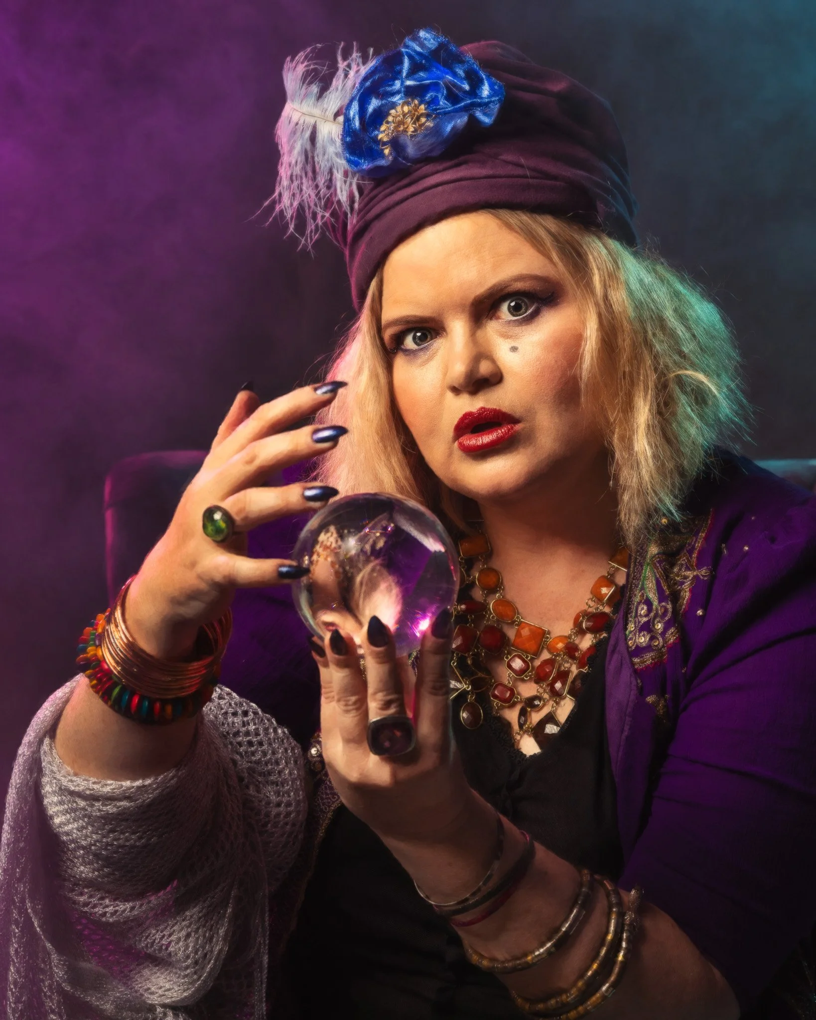 A blonde woman clad in purple robes and a purple turban holds a crystal ball and stares at the camera.