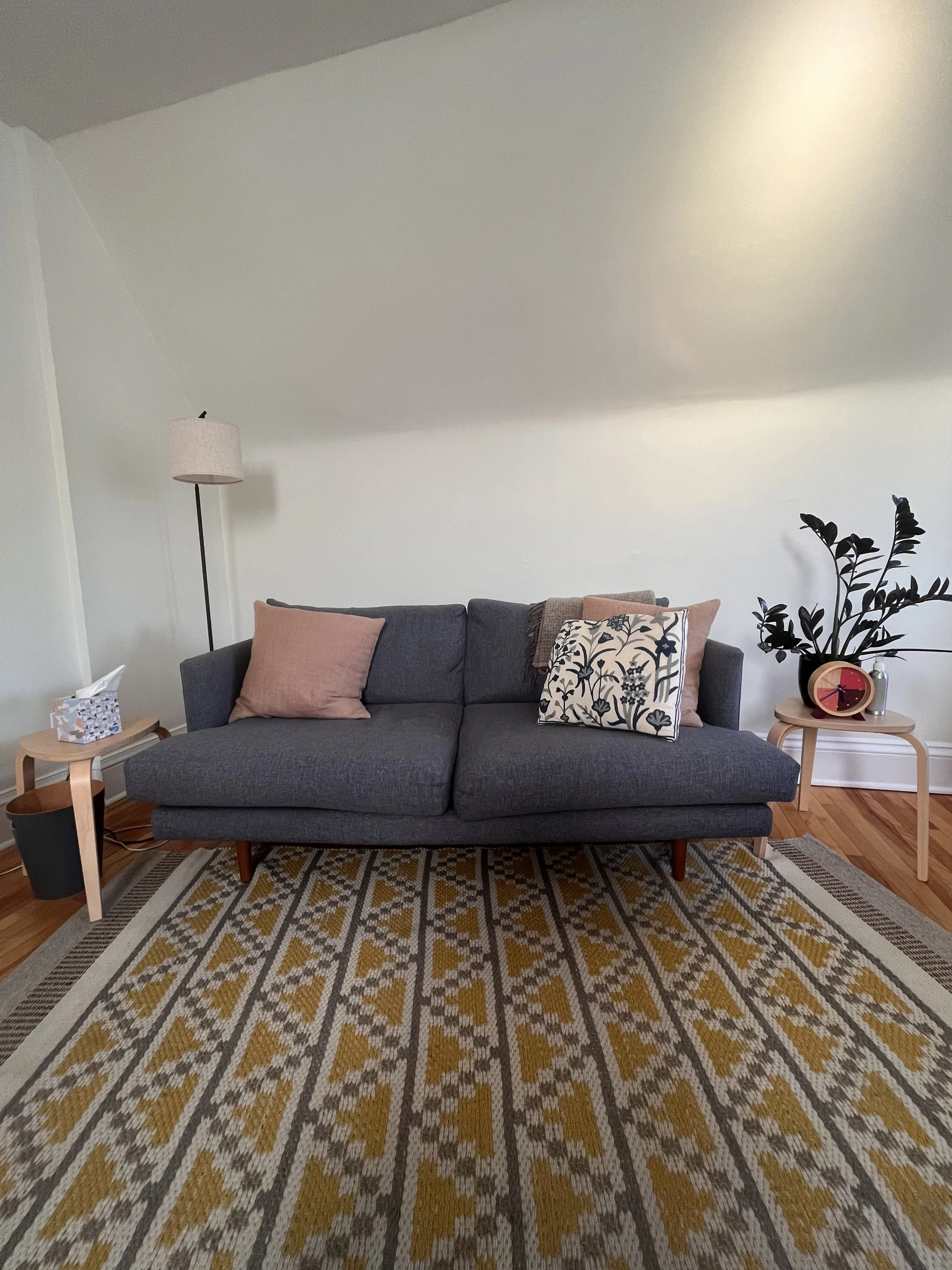 Therapy room with a dark gray sofa, pink and patterned pillows, a patterned area rug, side tables, a table clock, and a plant on a small table, with a white wall and ceiling in the background.
