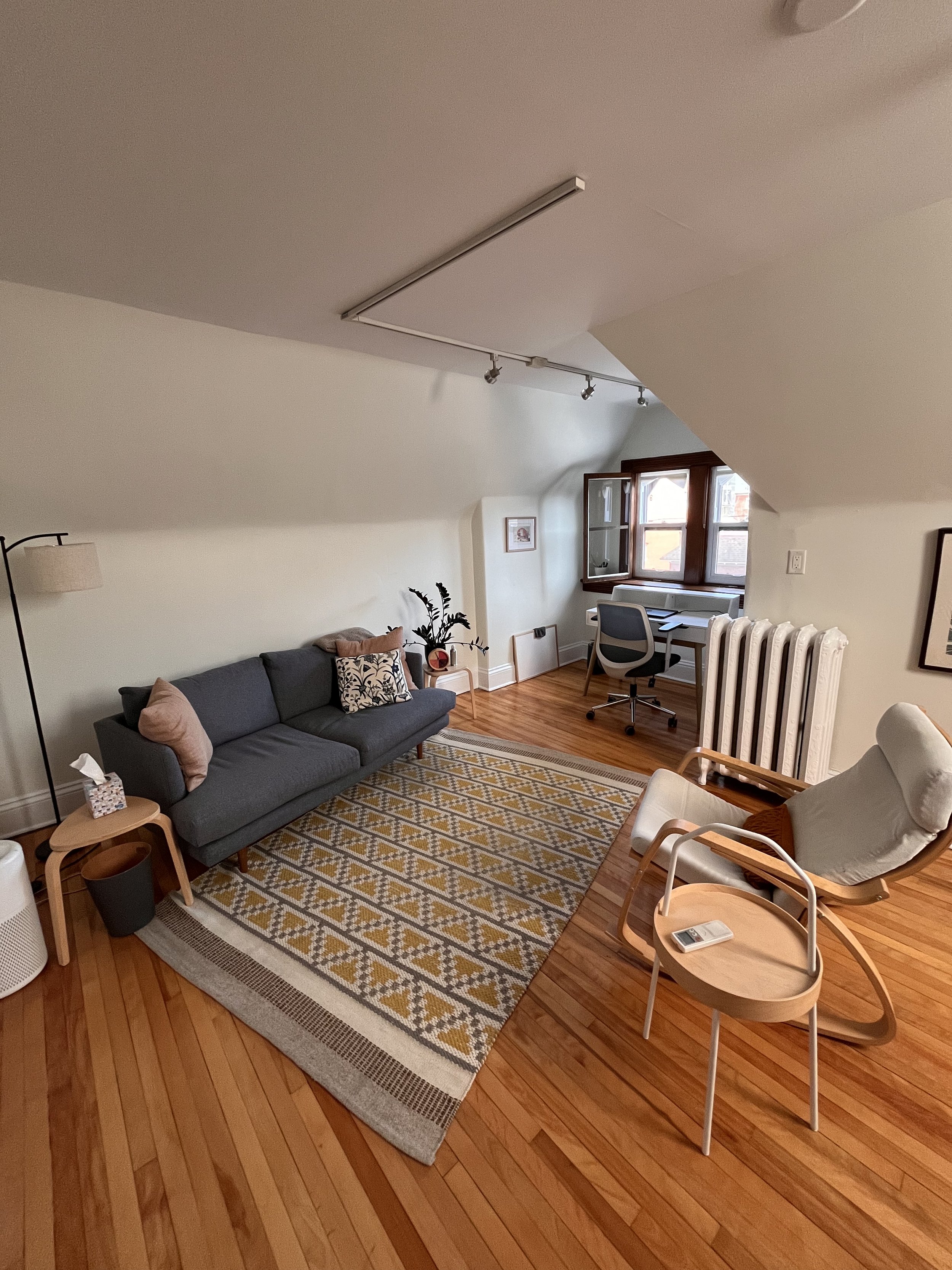 Therapy room with a dark gray sofa, patterned area rug, wooden floor, white walls, a side table with tissue box, a floor lamp, a small table near the sofa, a desk with an office chair near a window, a radiator, and a rocking chair with a pillow.