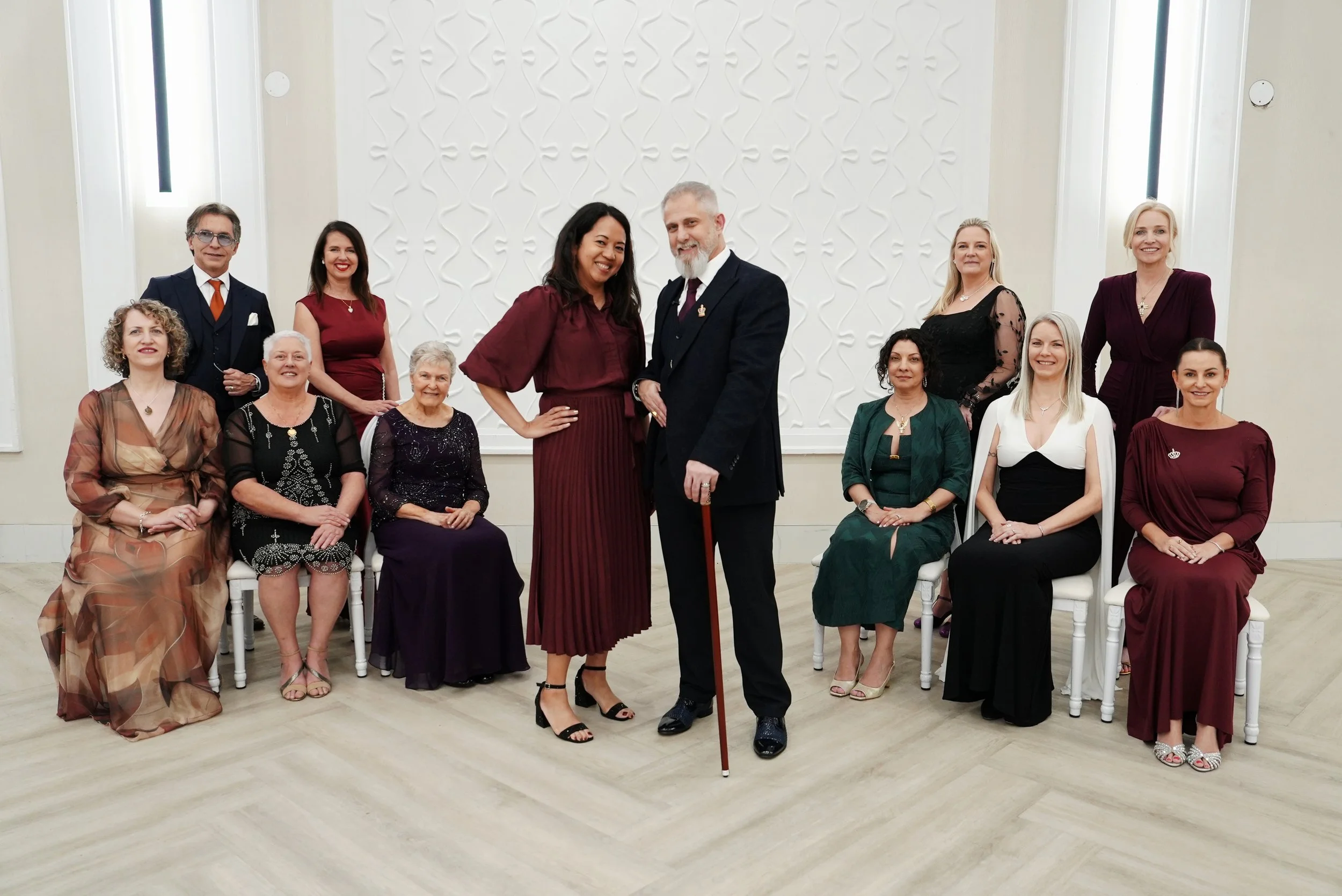 Group of people dressed formally, some seated and others standing, posing in a bright, modern room with a textured white wall background.