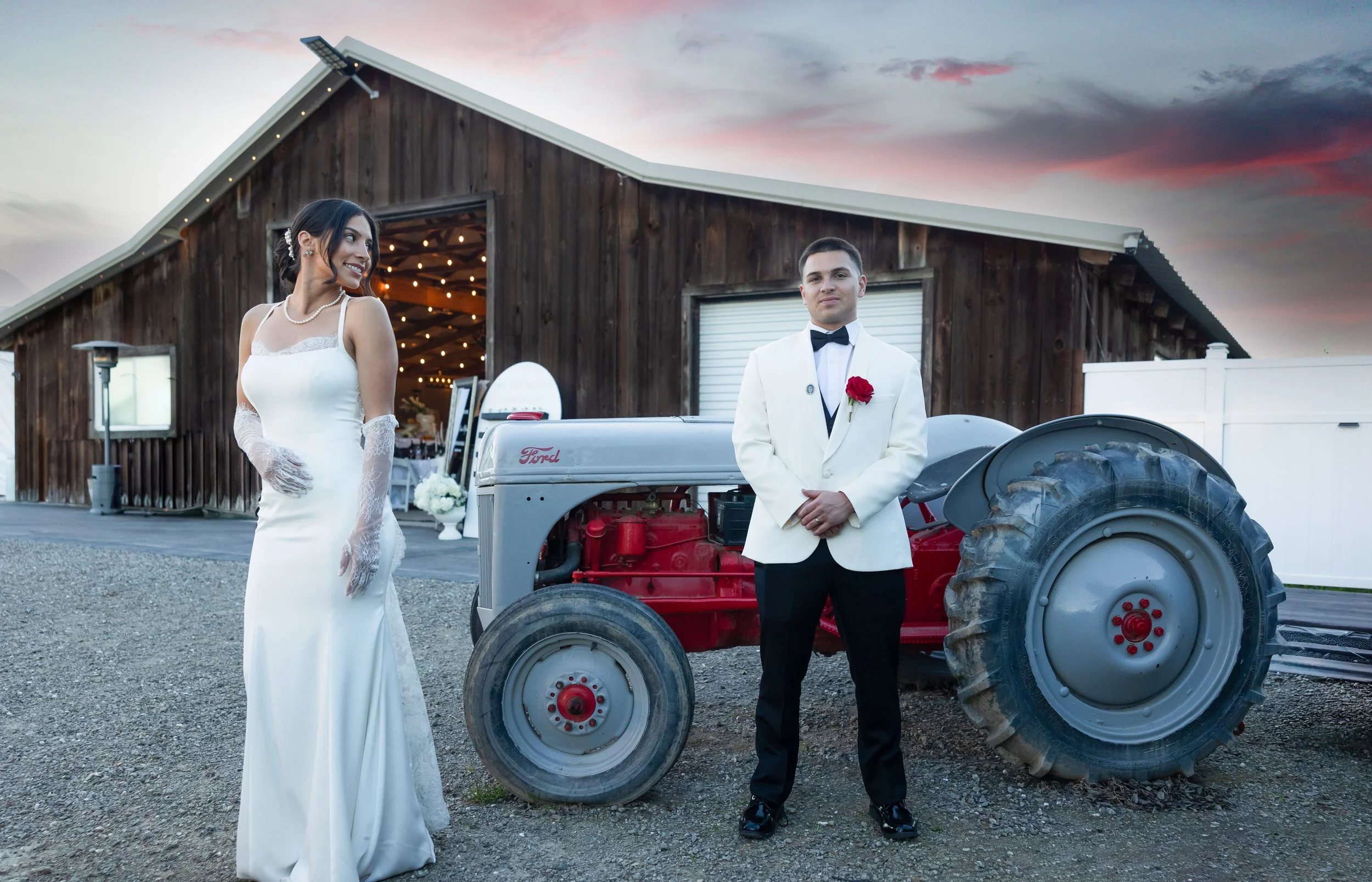 A bride and groom in wedding attire standing outdoors at sunset. The bride is in a white gown with lace gloves, and the groom is wearing a white tuxedo jacket with a red rose boutonniere. They are beside a vintage Ford tractor in front of a rustic wooden barn.