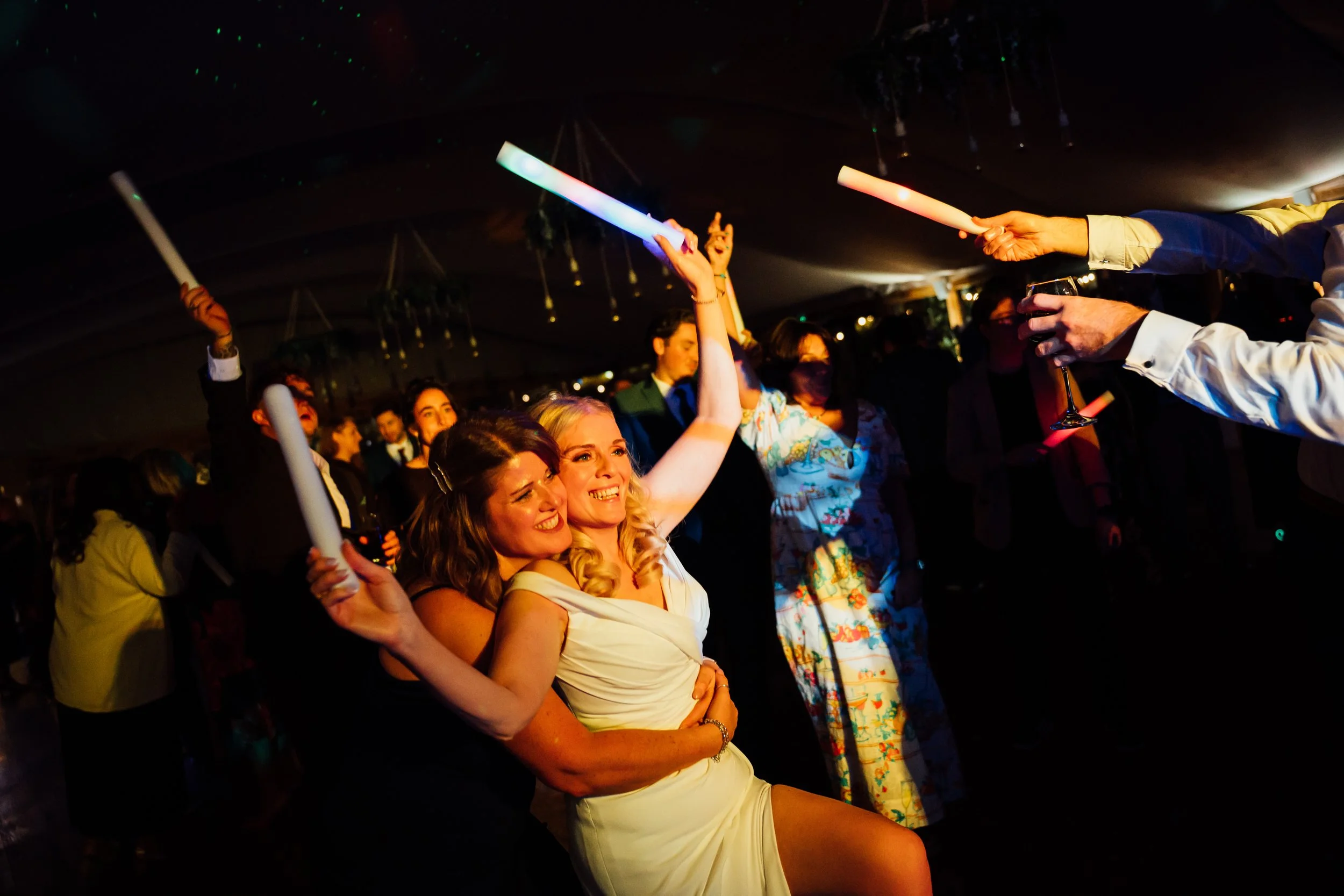 Group of people celebrating at a party, two women in front smiling and hugging, holding glow sticks, with more people dancing and conversing in the background.