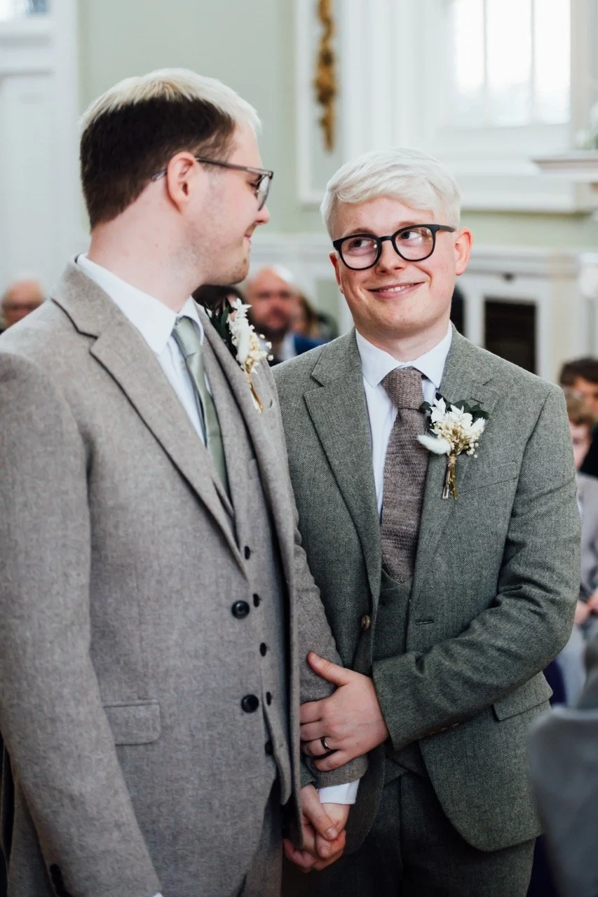 Two men, dressed in suits with floral boutonnières, hold hands at a wedding ceremony, exchanging vows or rings, with guests in the background.