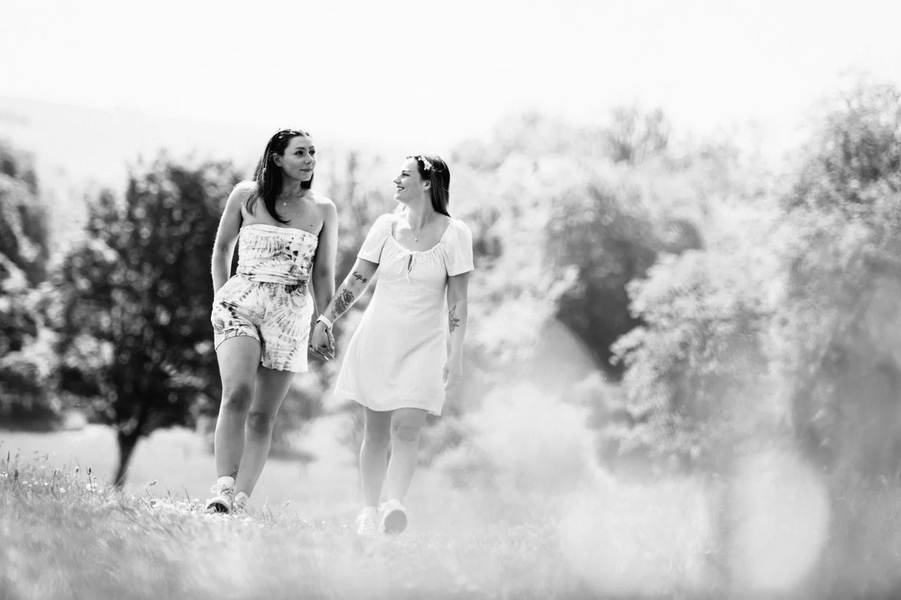 Two women walk hand-in-hand outdoors on a sunny day, surrounded by trees and grass in a park.