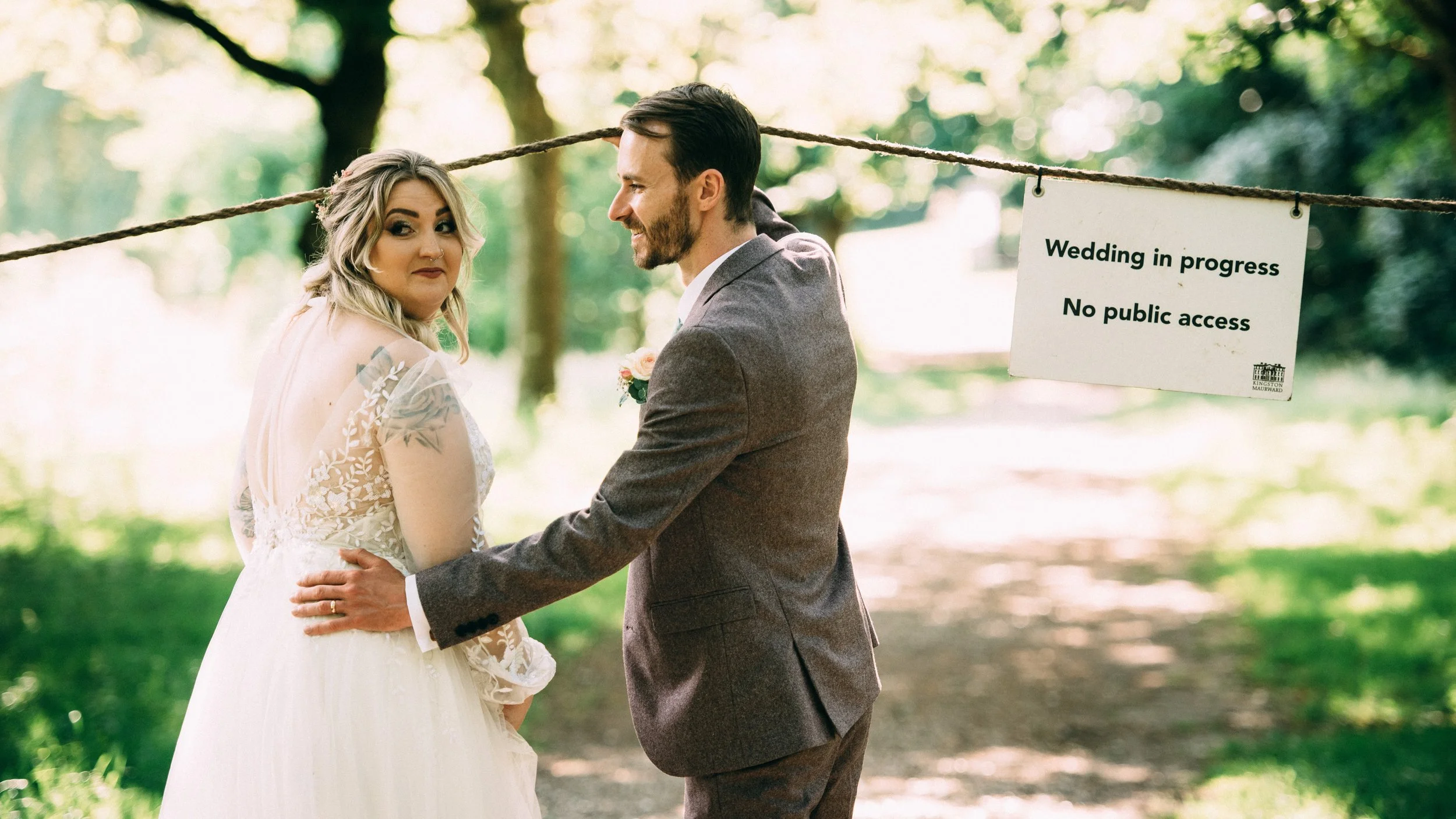 A bride and groom holding hands during an outdoor wedding in a wooded area, with a sign that reads "Wedding in progress, No public access" hanging from a rope.