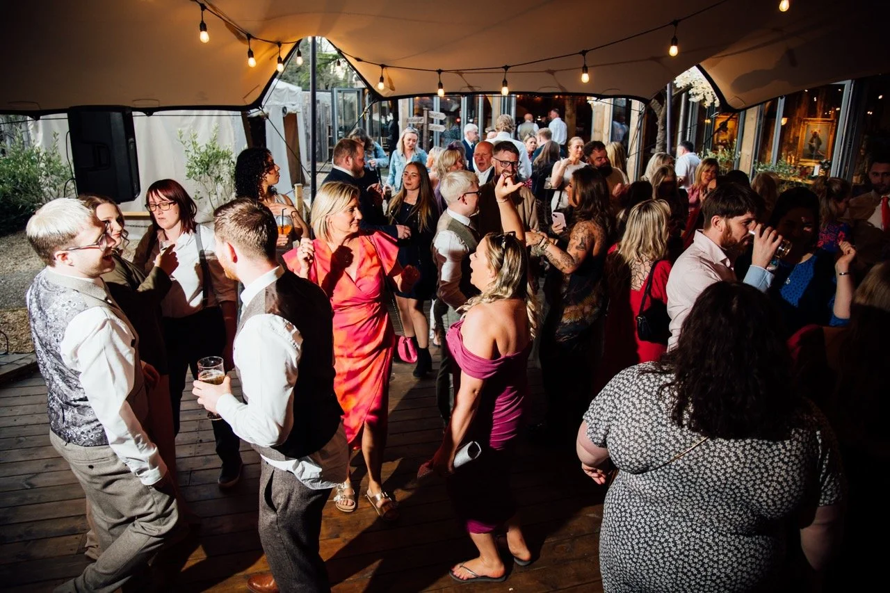 Group of people at a lively party dancing and socializing under string lights, with some holding drinks inside a decorated venue with large windows.