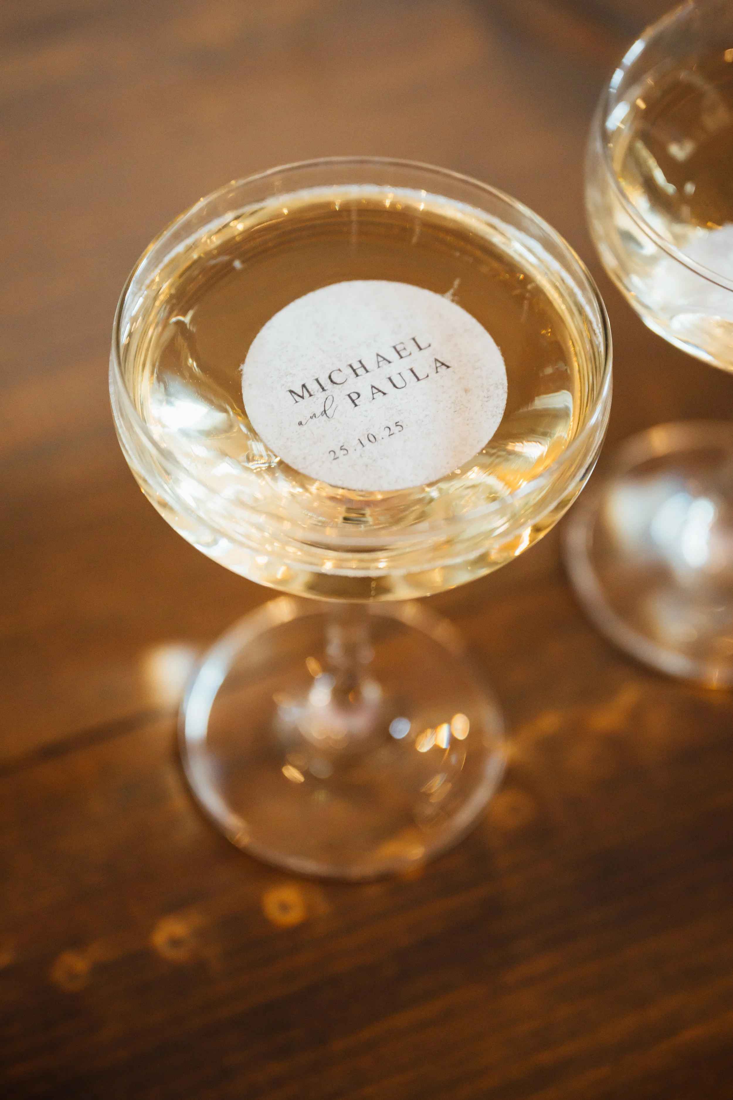 A wedding champagne glass with a custom sticker reading 'Michael and Paula 25.10.25' on the surface of the drink, with another similar glass partially visible beside it, on a wooden table.