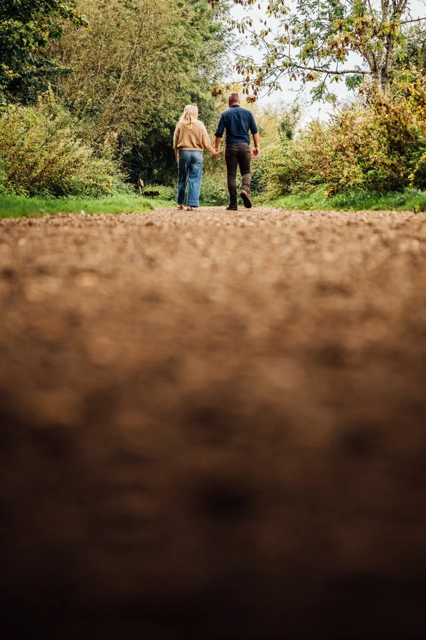 A couple walking hand in hand down a dirt path in a park with trees and bushes around them.